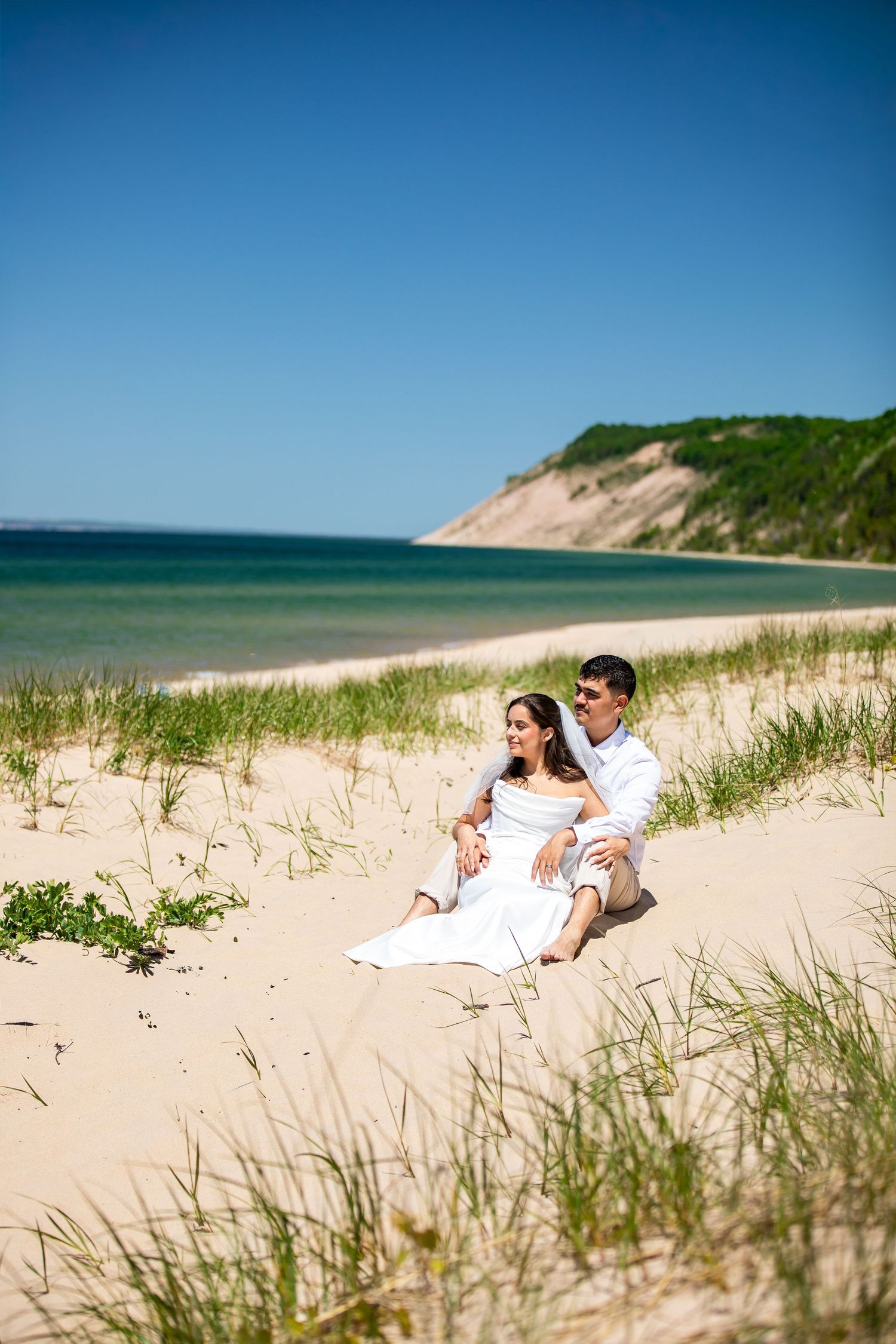 A couple in white attire sits romantically on sandy beach dunes with turquoise water and green hills in background.