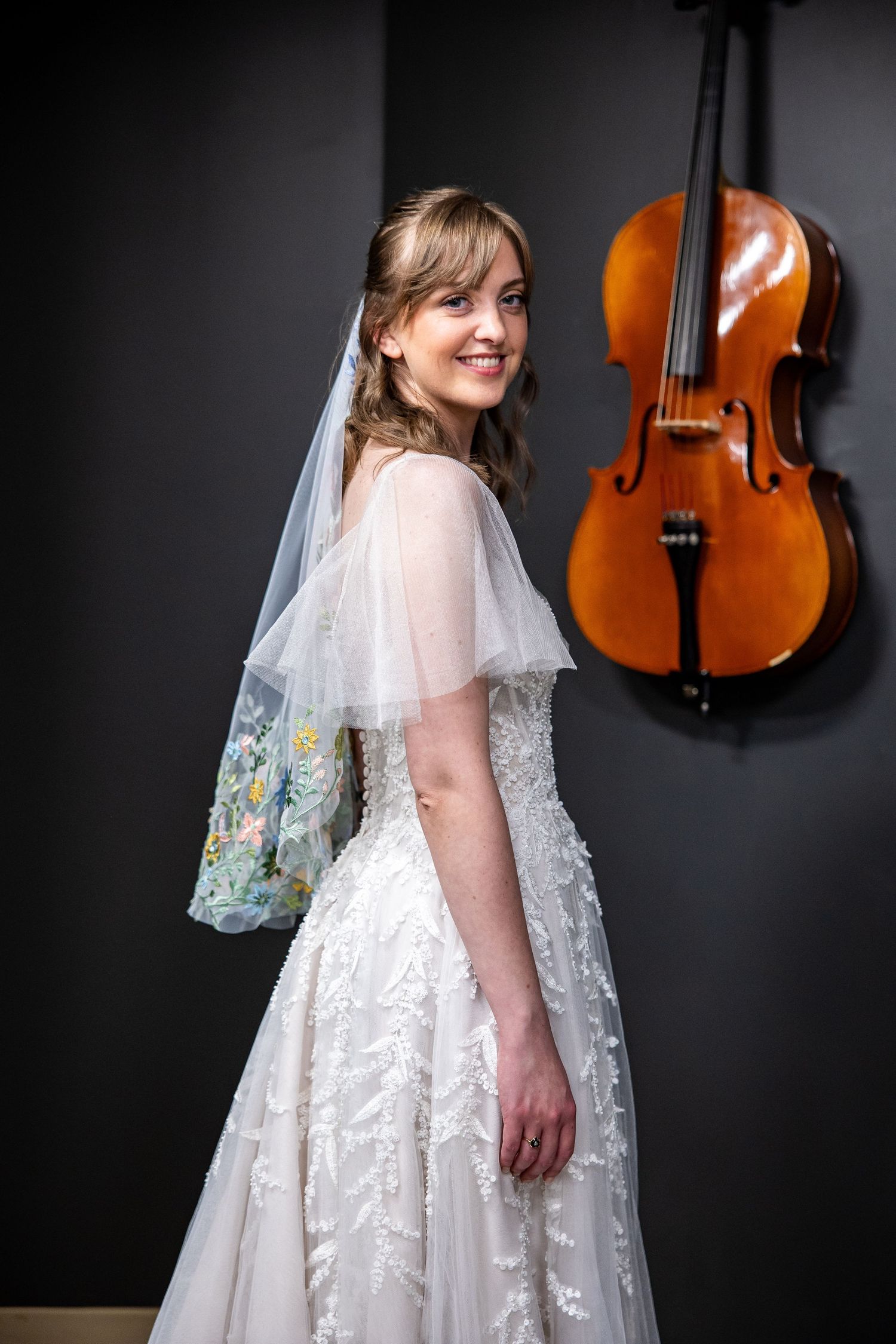 Smiling bride in white lace gown and floral veil poses beside a cello hanging on a dark gray wall.