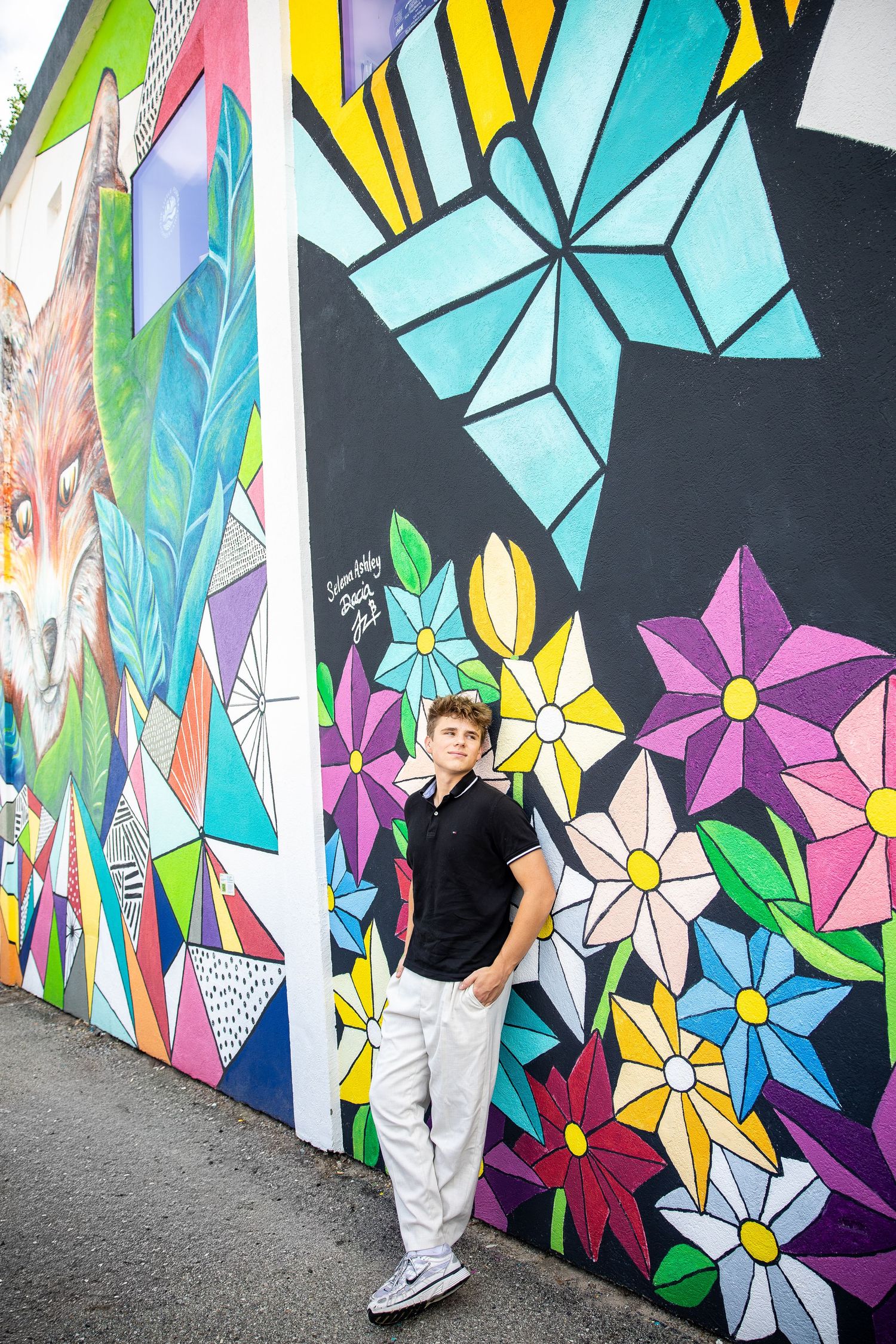 Teen boy in black polo and white pants poses confidently in front of a vibrant colorful floral street mural.