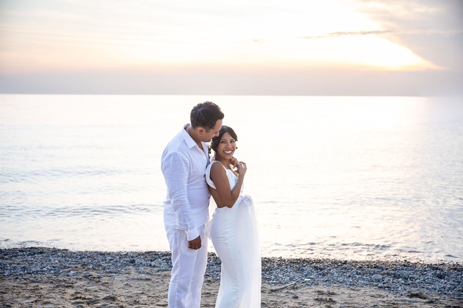 Bride and groom in white embrace on a pebbly beach at sunset, with calm sea and soft golden sky behind them.