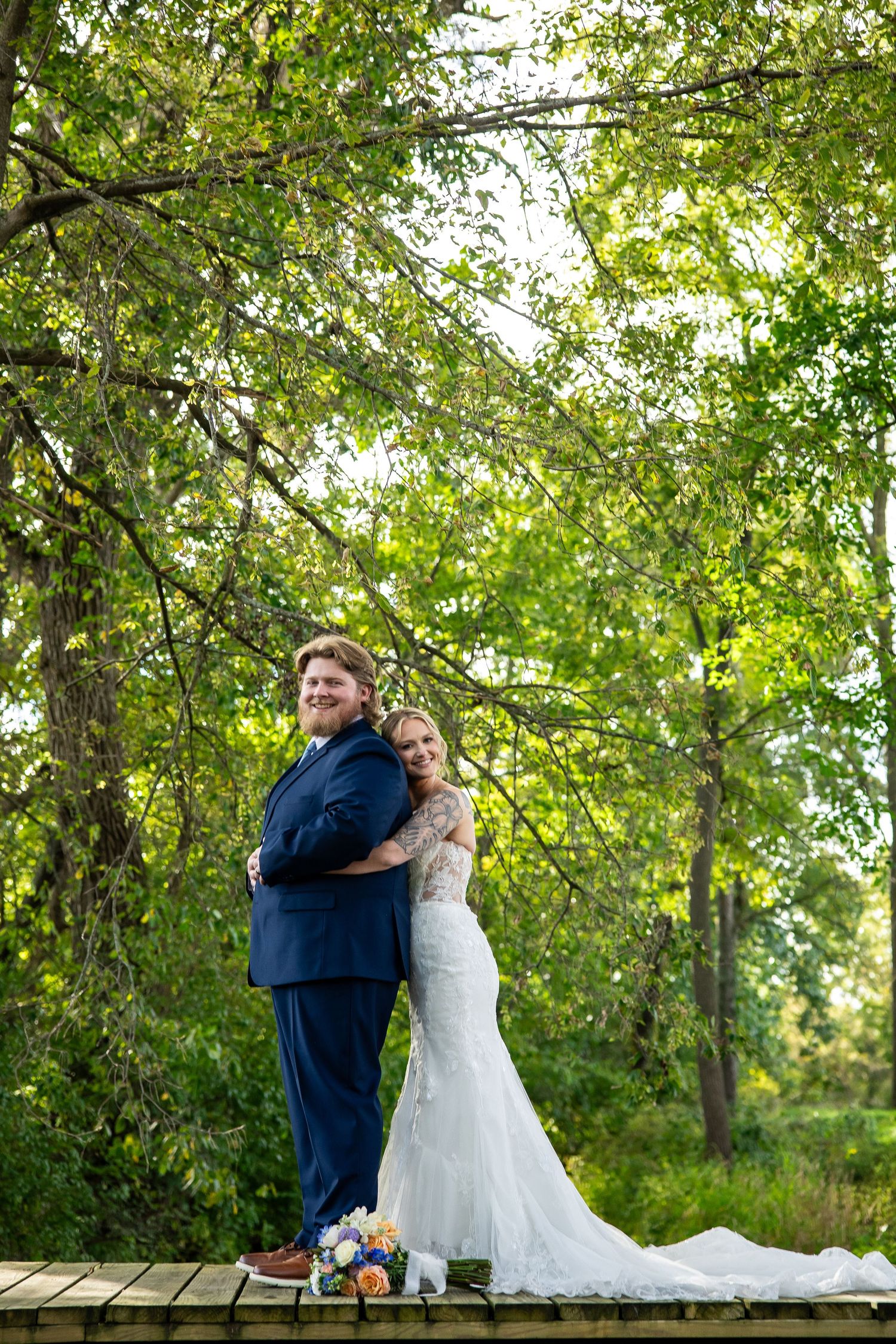 Bride in a white lace wedding gown and groom in a navy suit embrace back to back on a wooden bridge surrounded by lush green trees.