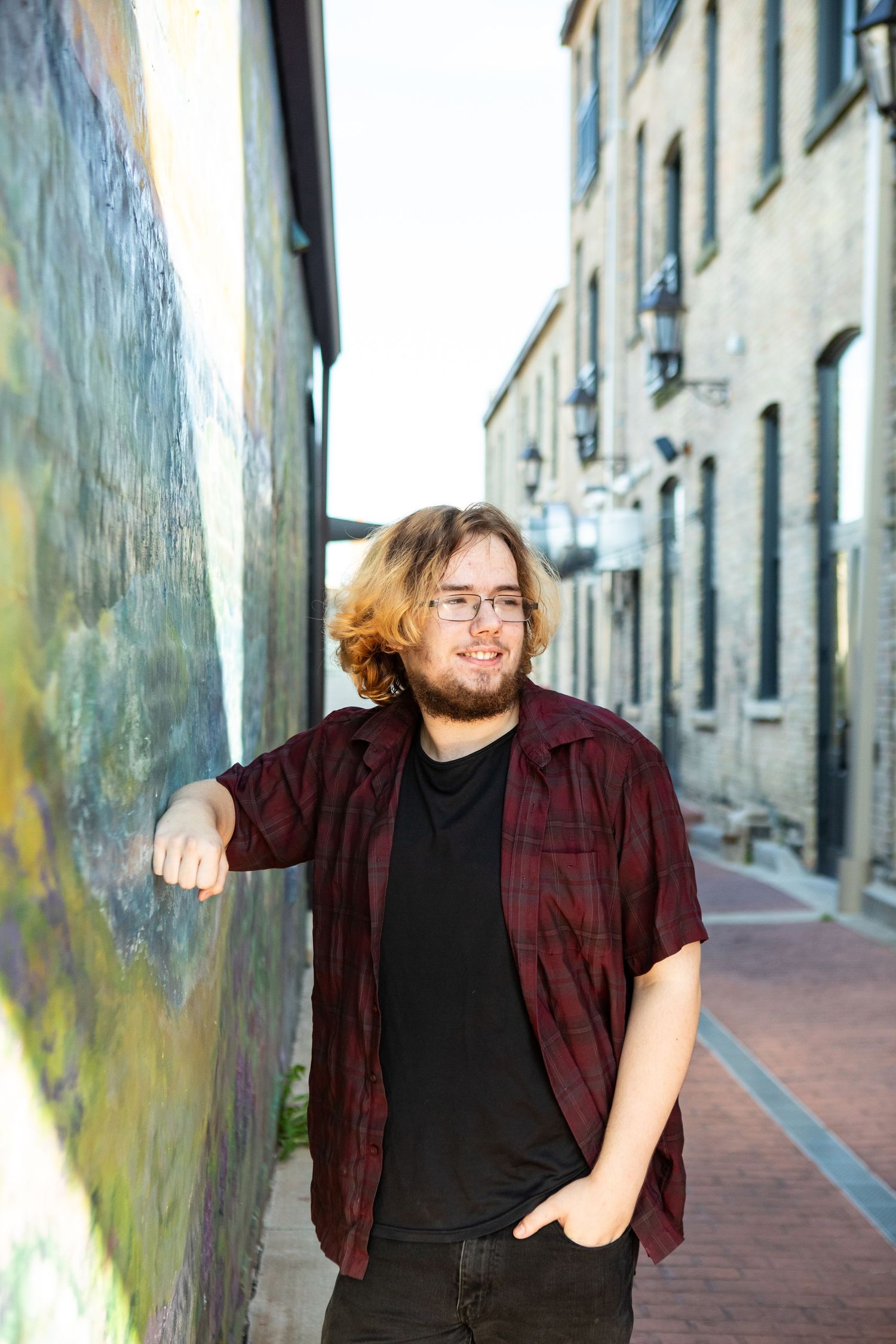 A person in a plaid shirt and glasses poses playfully against a stone wall along a narrow cobblestone street.