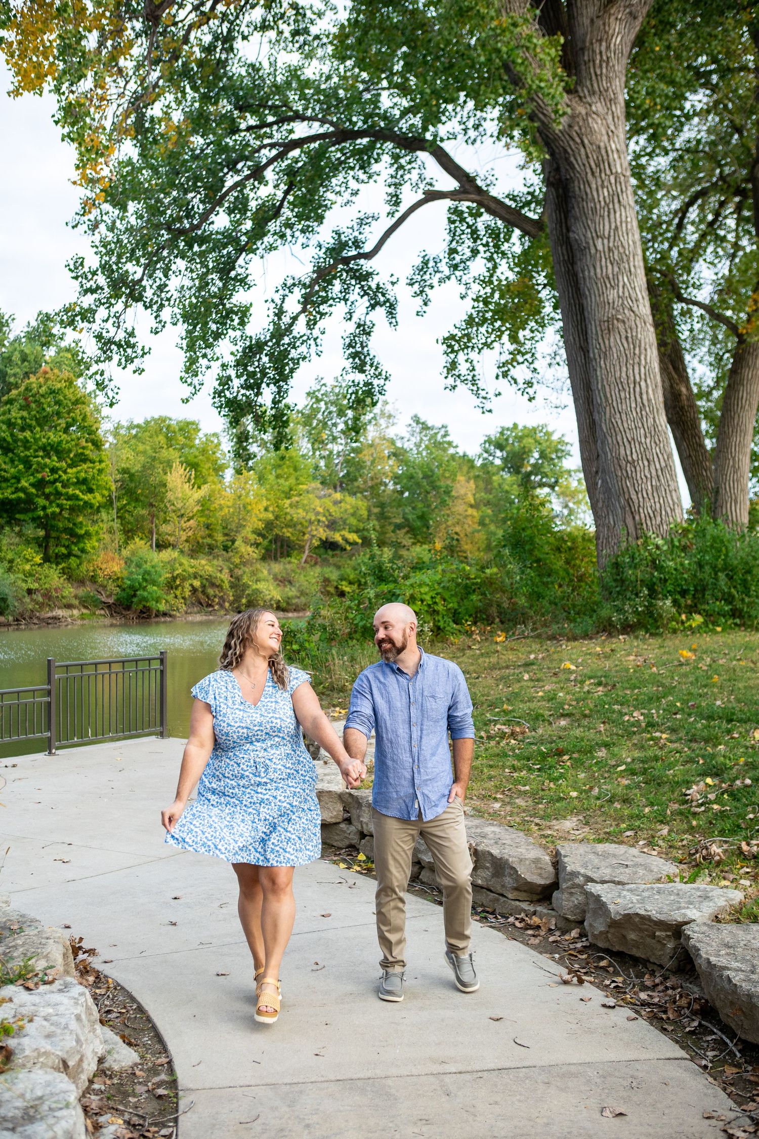 A couple walks together on a path by a lake surrounded by trees.