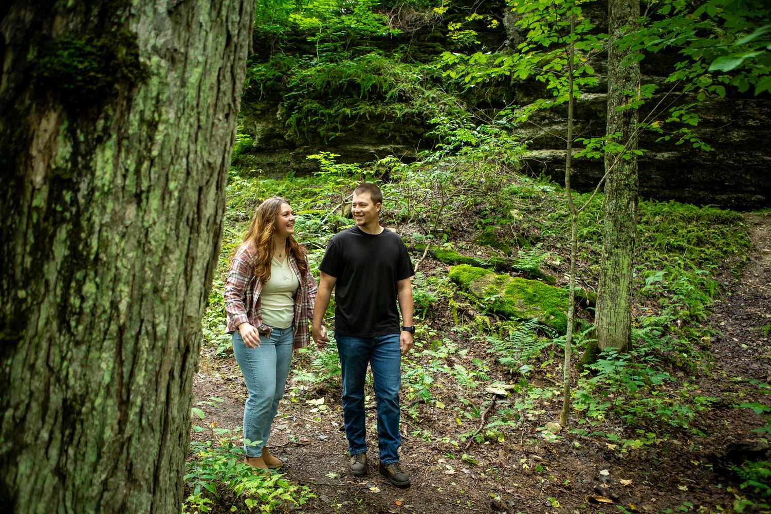 A couple walks through a wooded forest trail holding hands.