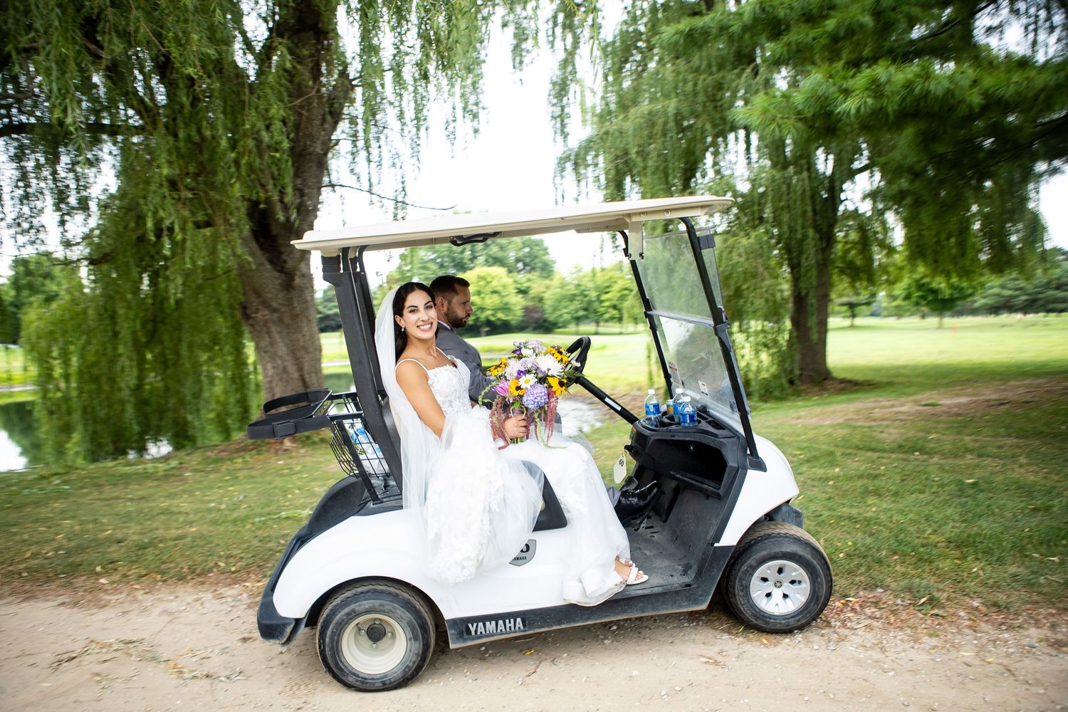 Bride and groom sitting together in a Yamaha golf cart on a scenic golf course with weeping willow trees.