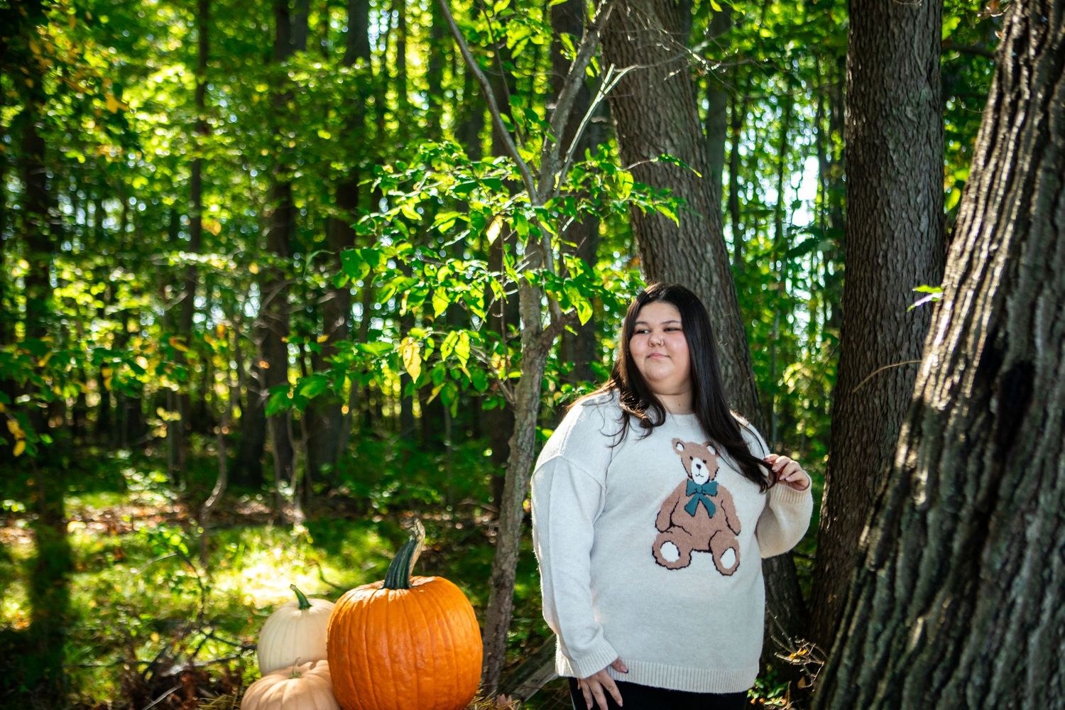 Woman in bear sweatshirt poses by forest trees with orange pumpkins in autumn outdoor photo session.