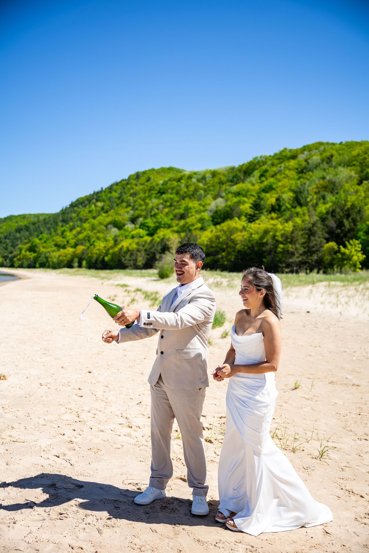 Newlyweds celebrate on a sandy beach, groom popping champagne in a tan suit beside bride in white gown.