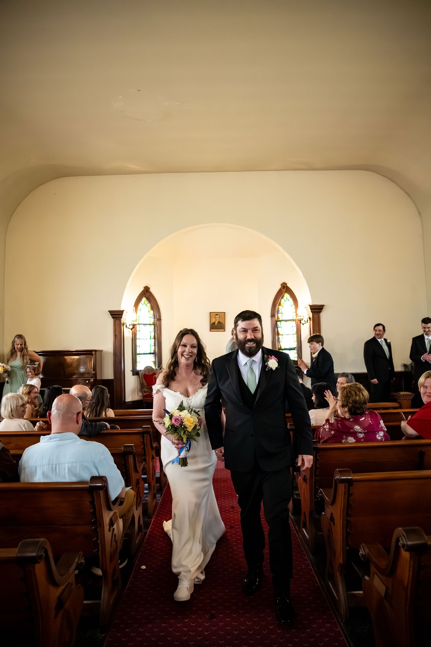 Bride and groom walk hand-in-hand down church aisle after wedding ceremony, bride holding colorful bouquet.