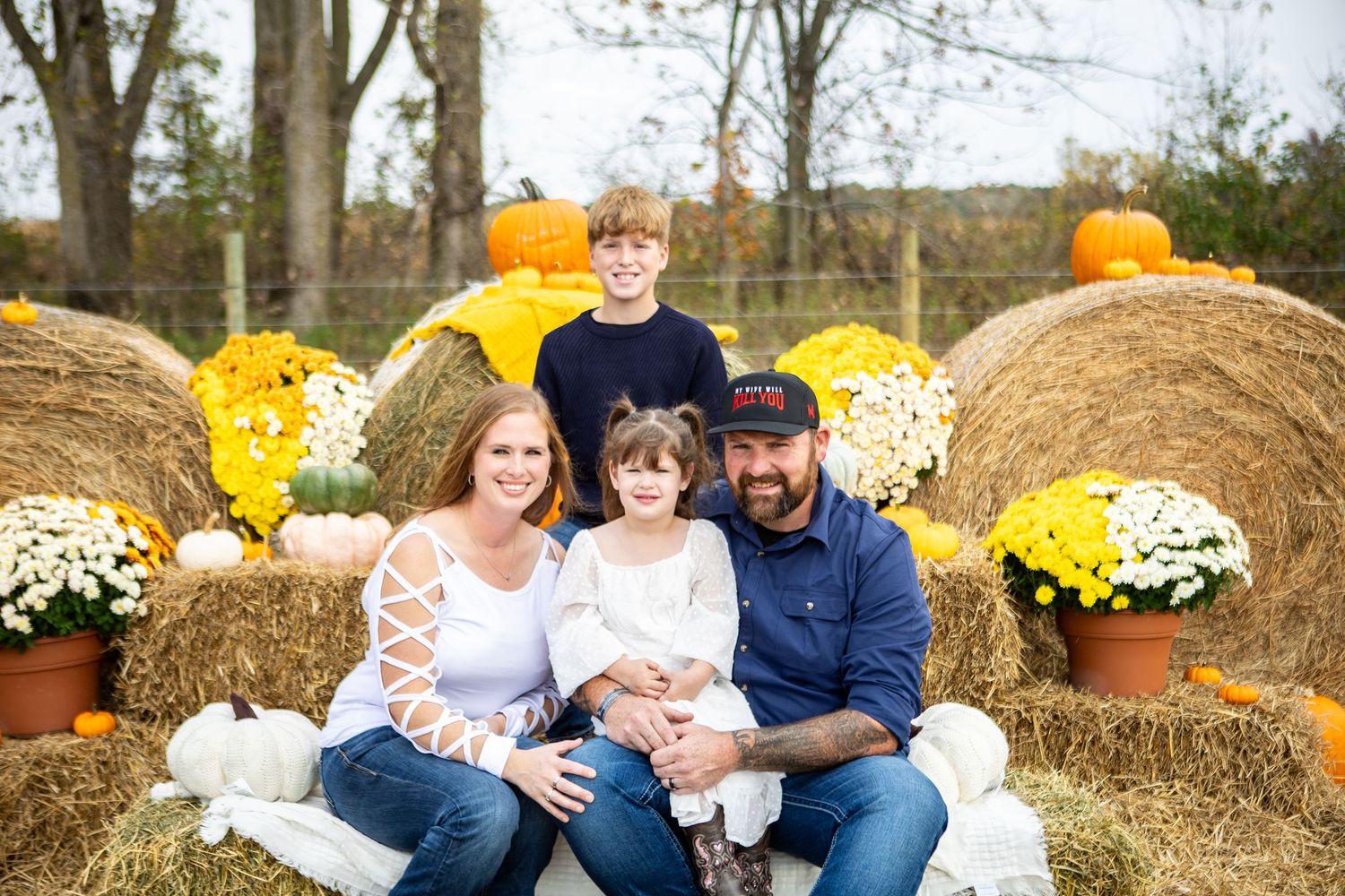 Family sitting together on hay bales surrounded by bright yellow mums and fall decorations in an outdoor setting.