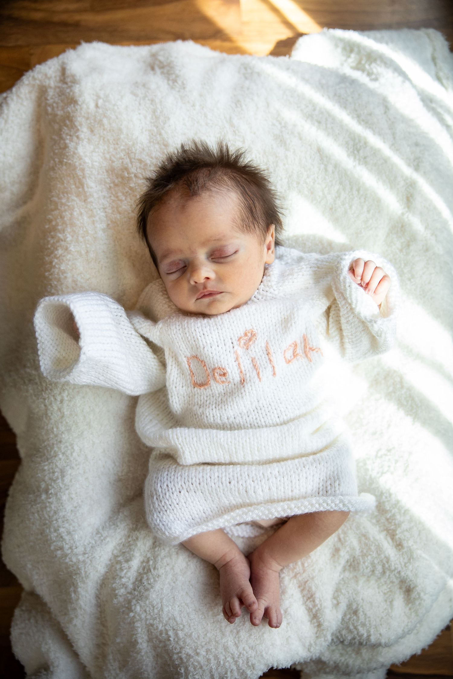 Sleeping newborn baby in white knit sweater with name embroidered, lying on soft white blanket.