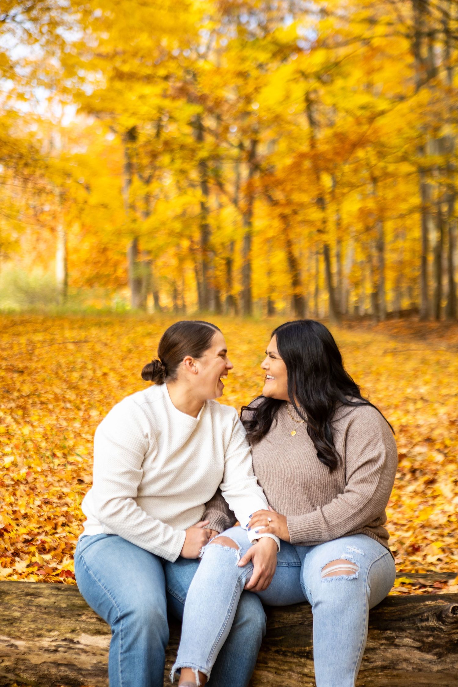 Two women sitting on a log in an autumn forest, smiling at each other, wearing cozy sweaters and ripped jeans.