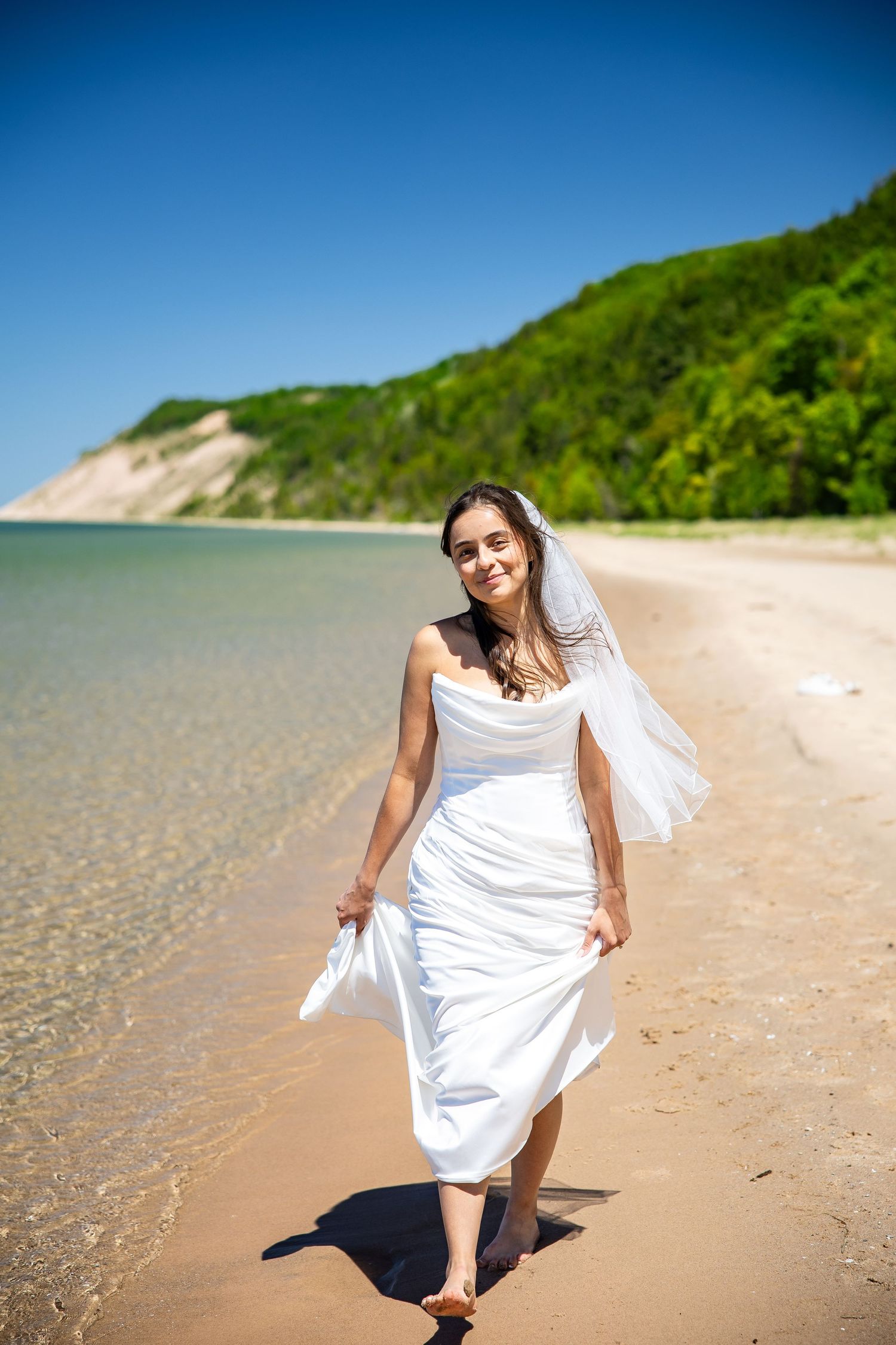 Bride in white wedding dress and veil walking barefoot on sandy beach with clear blue water and green hills.