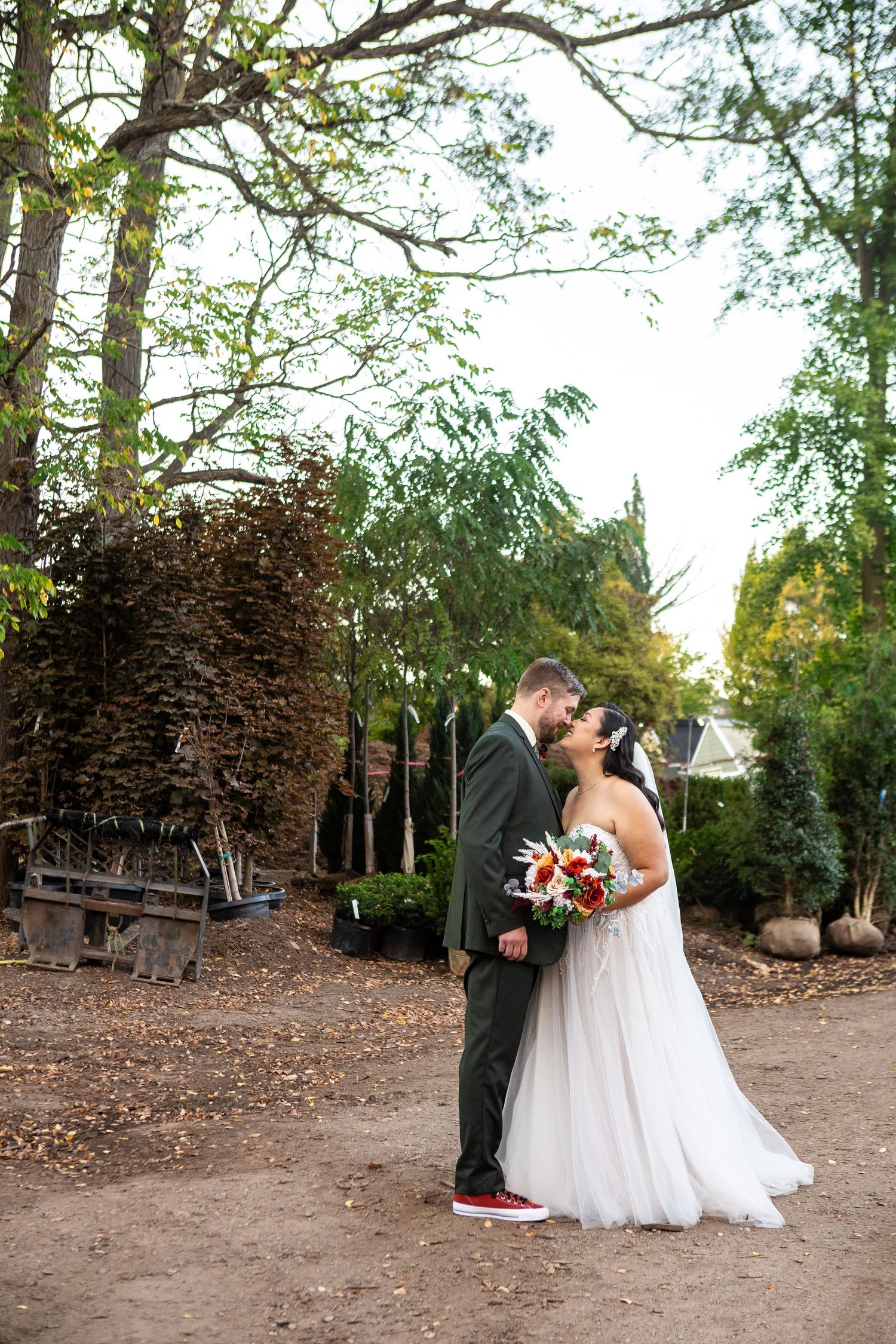 Bride and groom share a kiss outdoors, she holds a colorful bouquet, surrounded by lush trees at their wedding venue.