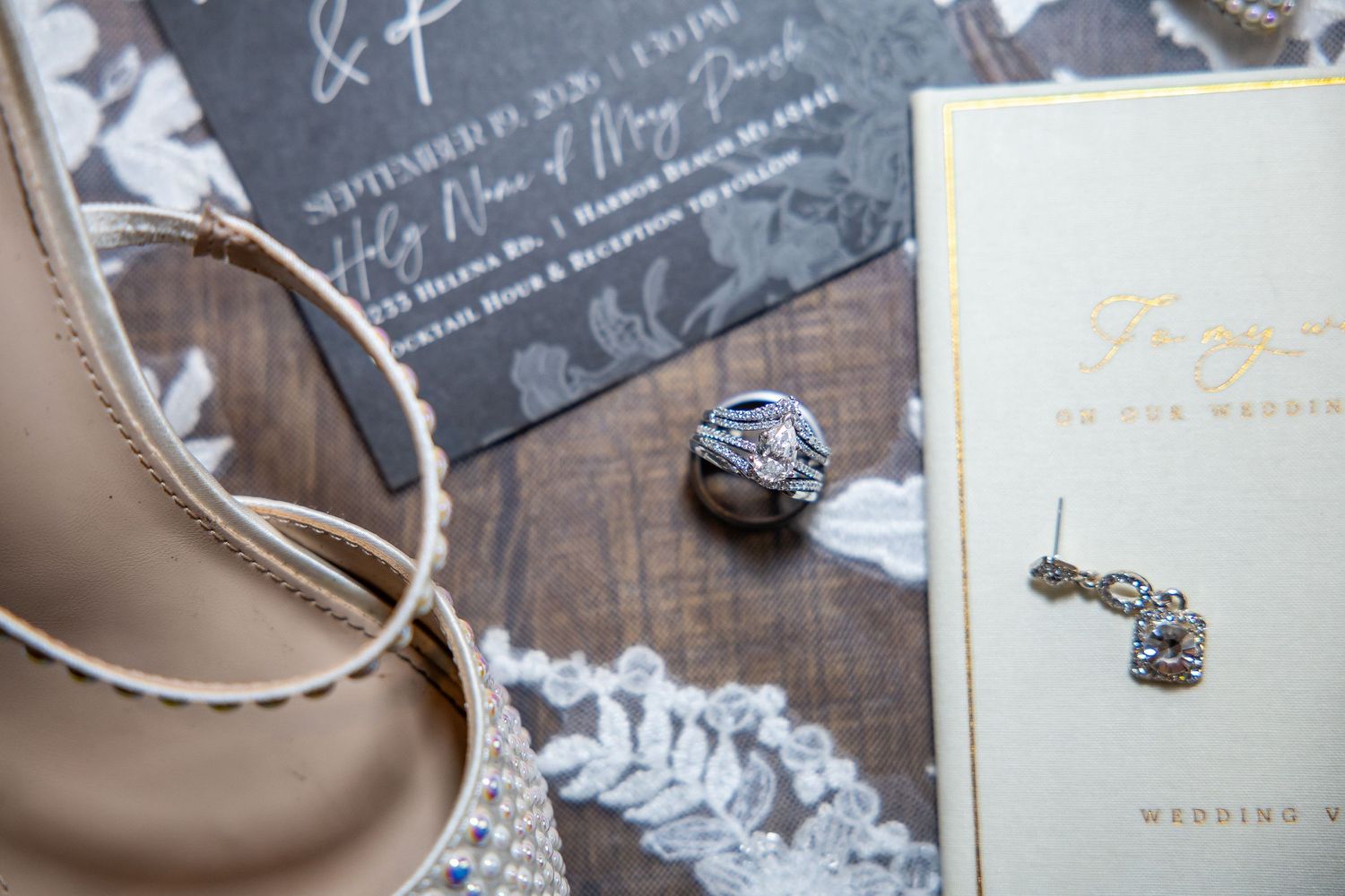 Wedding flat lay with a beige strappy heel, lace fabric, dangling crystal earrings, a ring and elegant invitation cards on a wood surface.