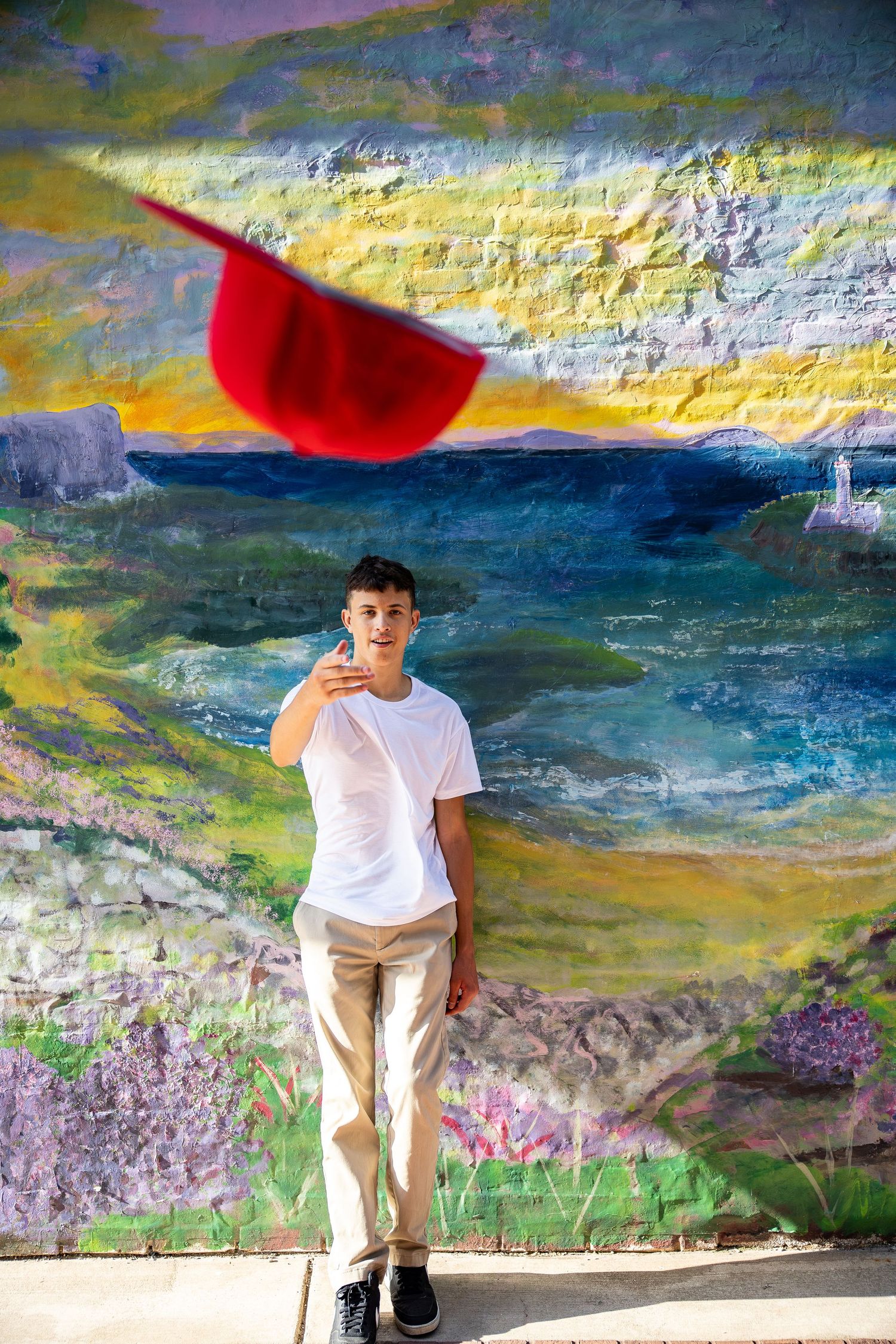 Teen boy in white tee poses in front of colorful landscape mural with red hat flying in air.