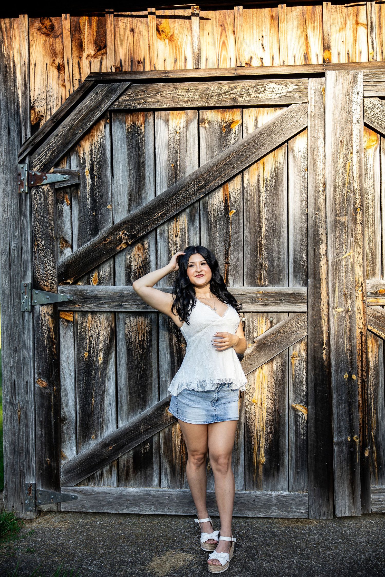 Young woman in white top and denim shorts poses confidently in front of rustic wooden barn doors in warm sunlight.