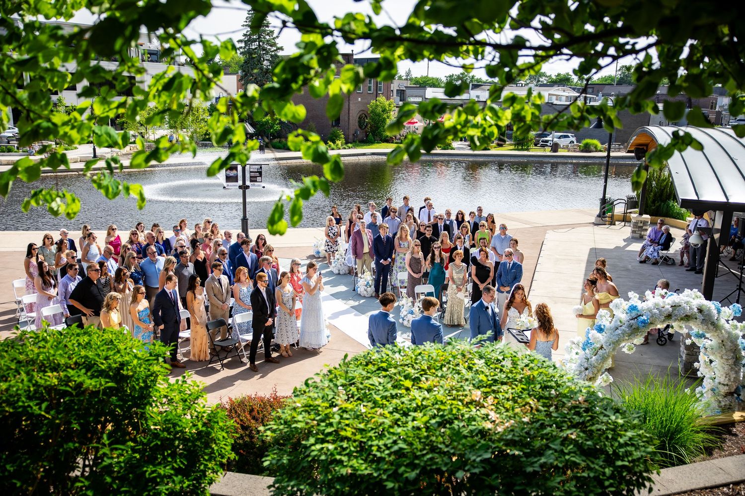 Outdoor wedding ceremony by a waterfront with guests gathered around a floral arch on a sunny day.