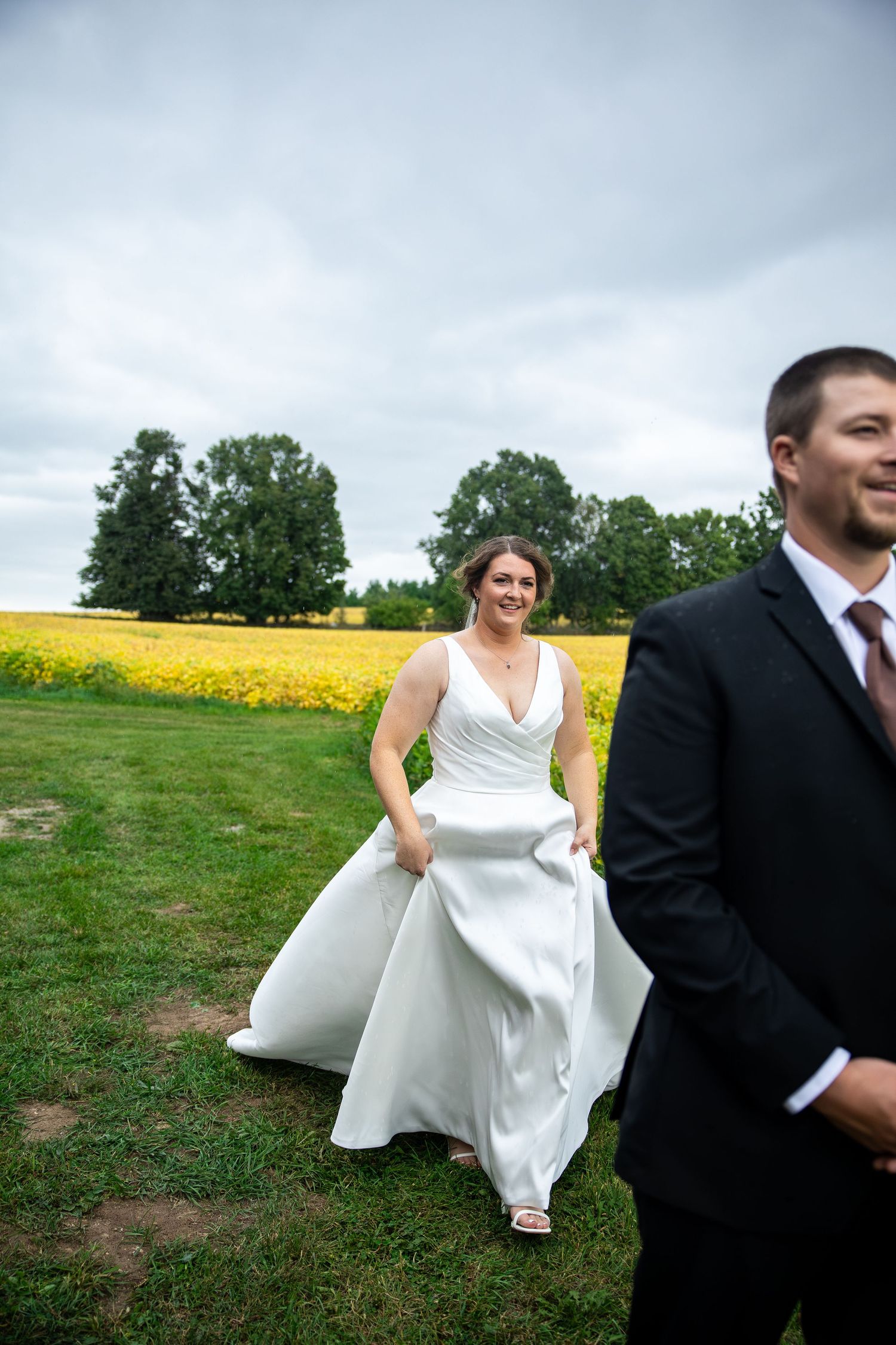 Bride in white gown laughs while walking toward groom in black suit near yellow flower field on cloudy wedding day.