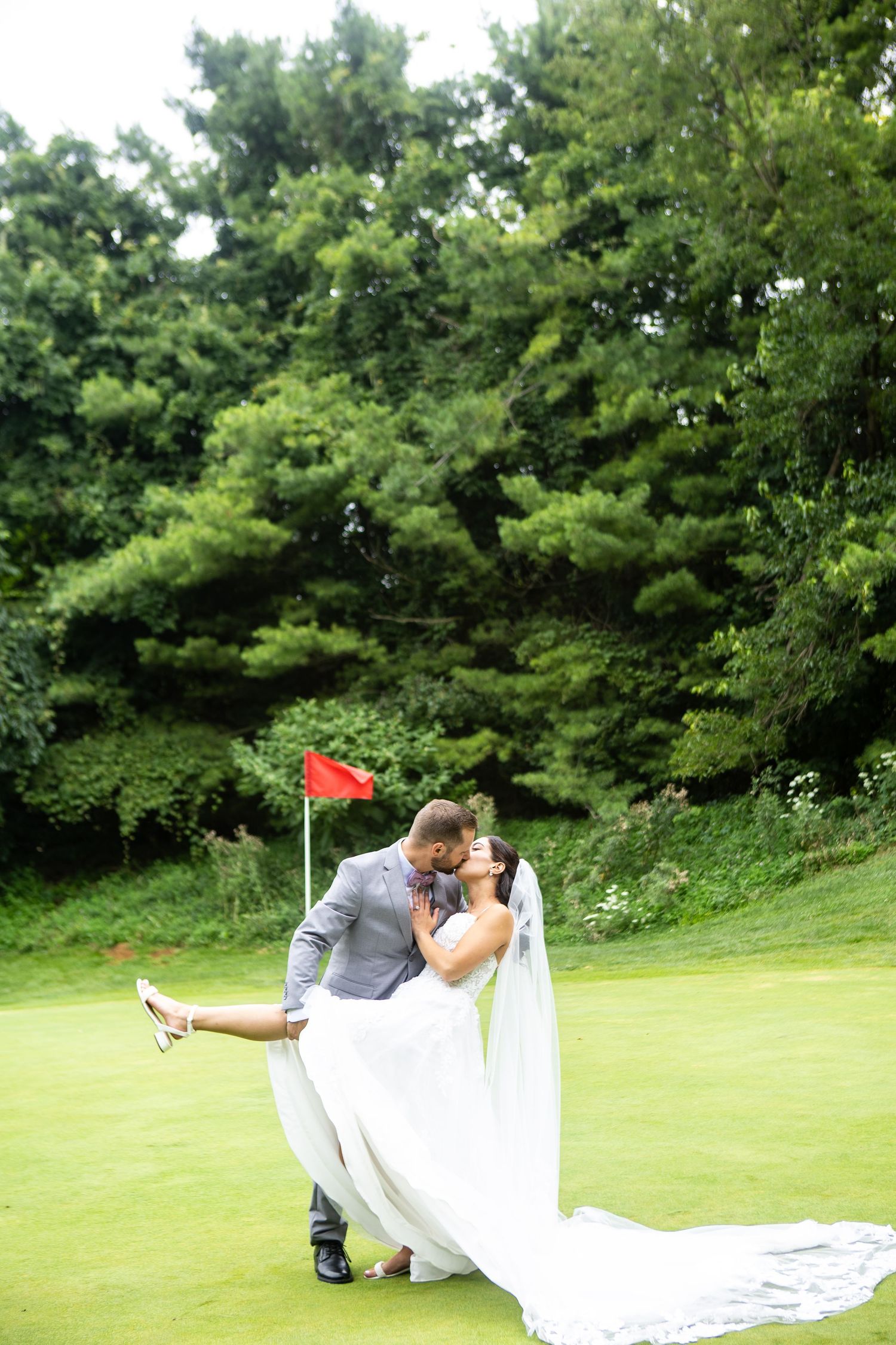 Groom dips bride in white gown for a kiss on a golf course green, with lush trees and red flag in background.
