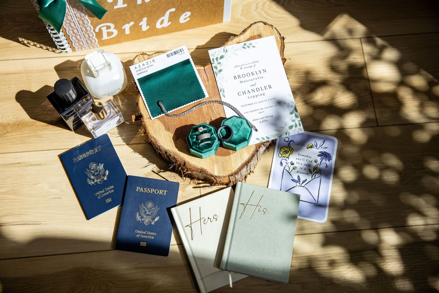 Flat lay of wedding details with navy passports, green vow books, a wedding invitation, and emerald accessories on a rustic wood table.