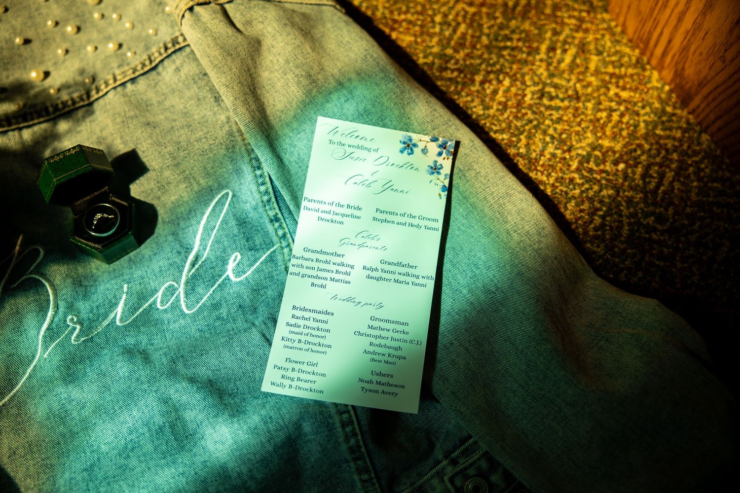 Denim bride jacket with white script lettering lies beside a green ring box and a floral wedding program card on a sunlit textured surface.