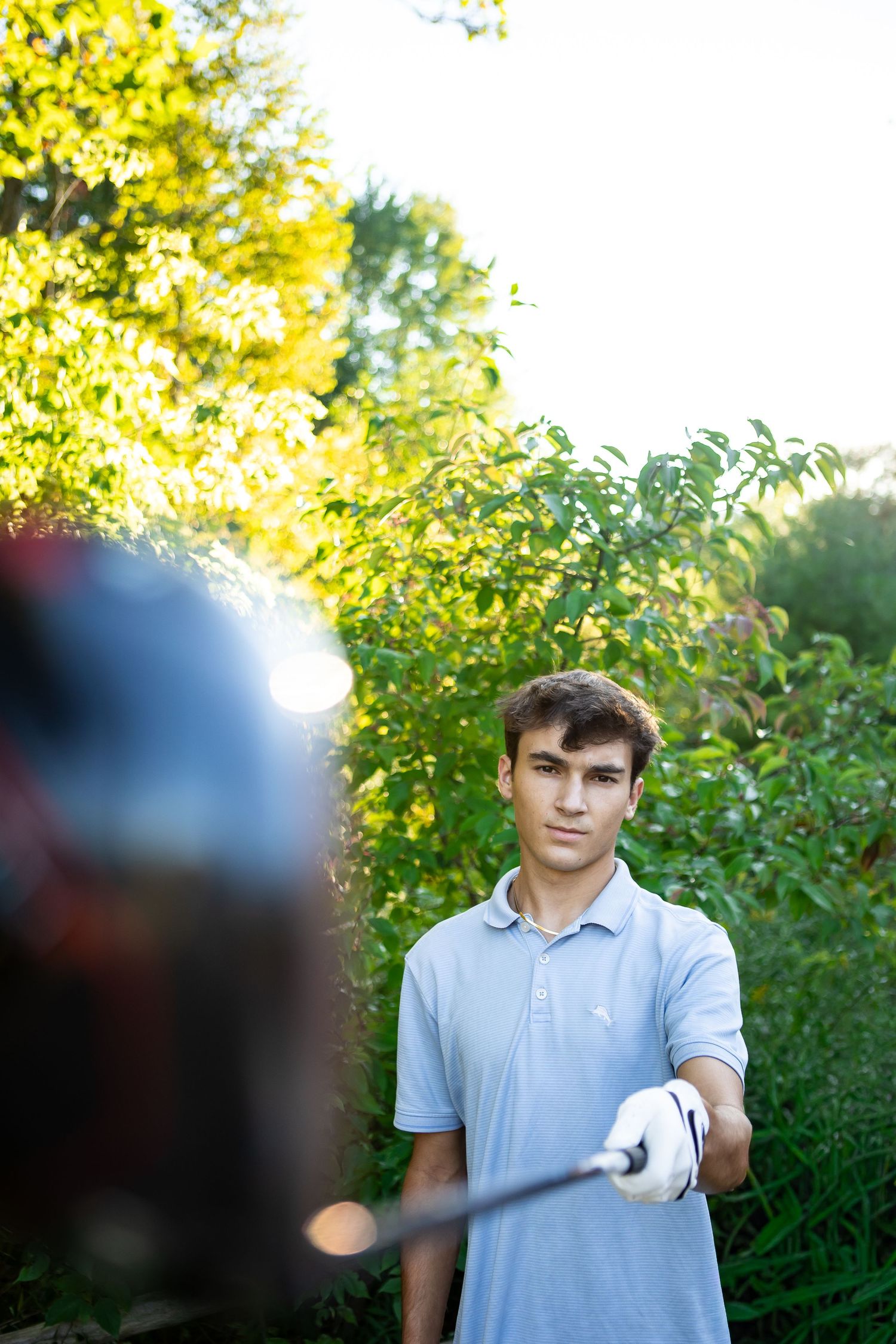 A person wearing a light blue polo shirt stands in front of lush green foliage on a sunny day.