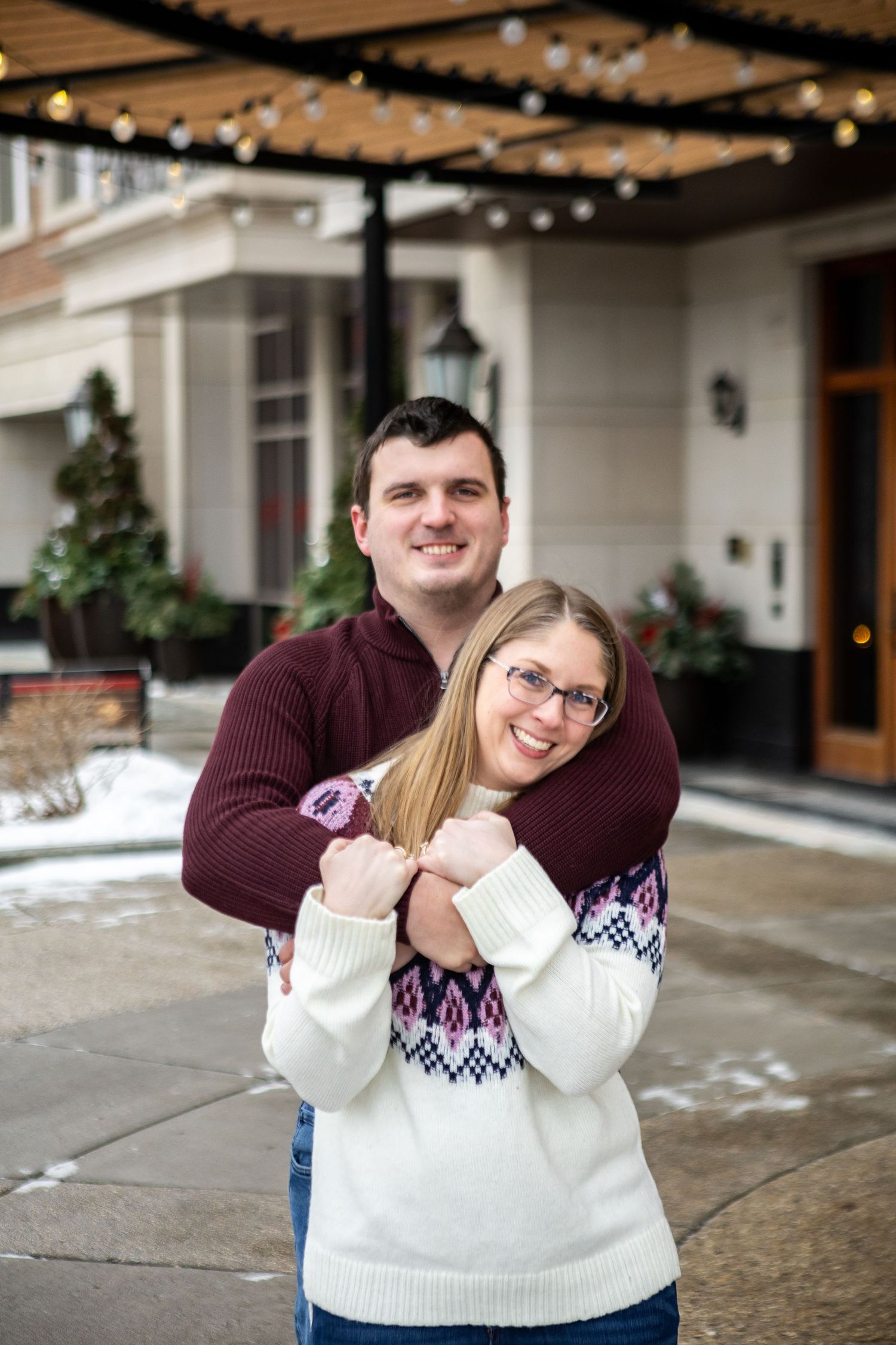 A couple wearing burgundy and white sweaters embrace lovingly on a snowy winter day outside a building.