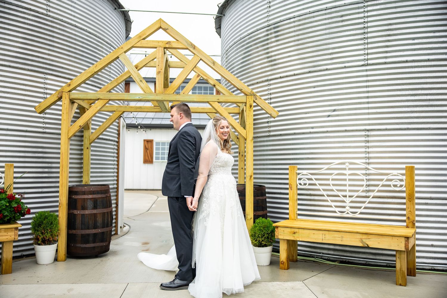 Bride in a white wedding gown and groom in a dark suit stand hand in hand beside a wooden gazebo, barrels and corrugated metal silos.