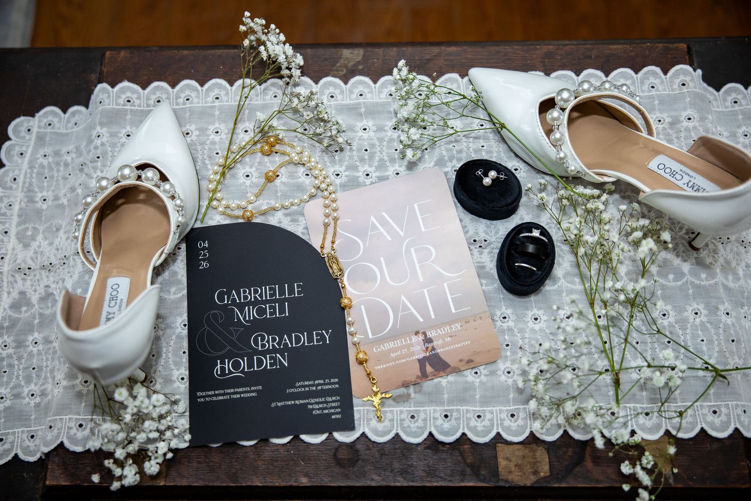 White bridal heels, gold jewelry, save the date cards, sunglasses, and baby's breath flowers arranged on a lace table runner.