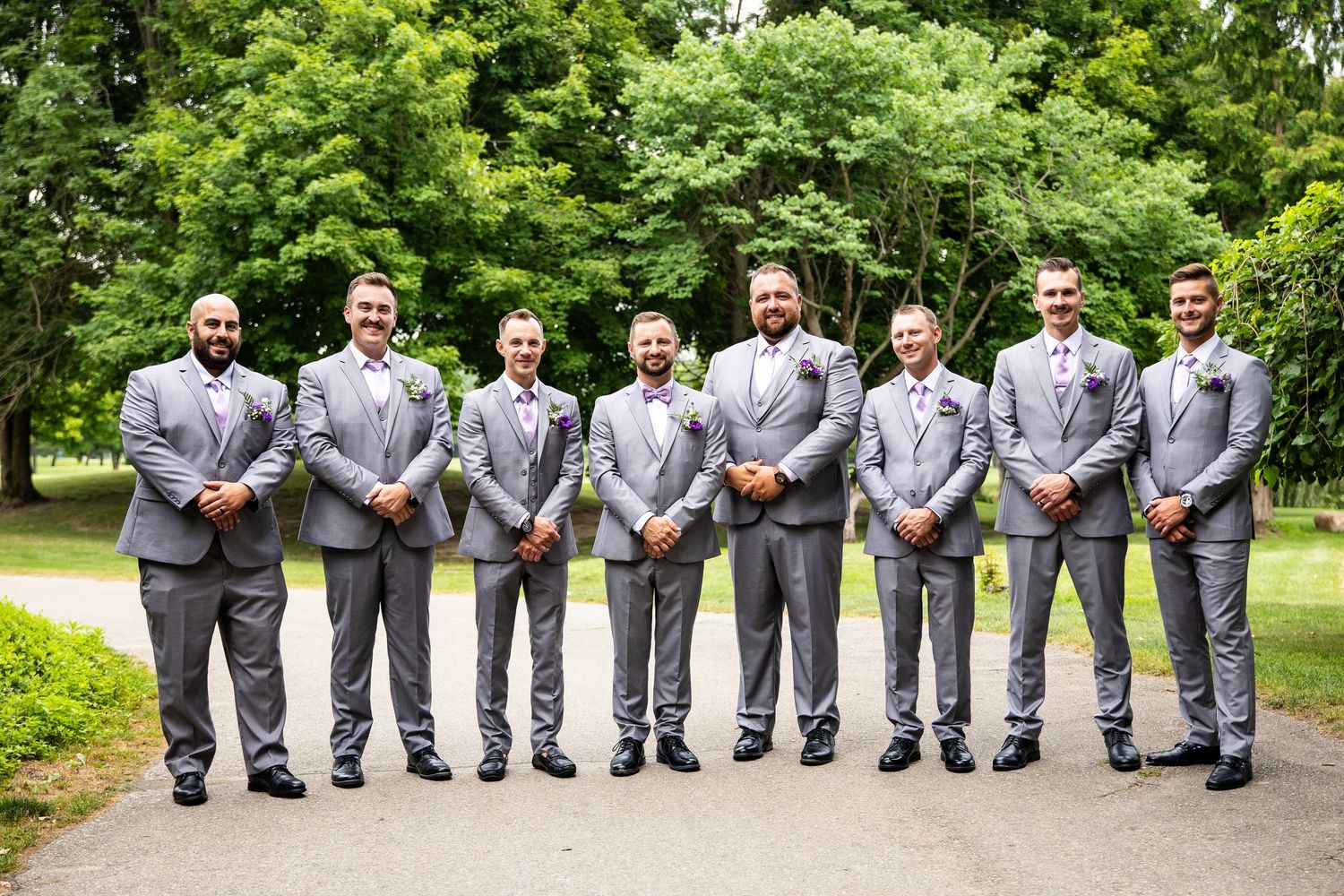 Eight groomsmen in matching gray suits with pink boutonnieres pose outdoors on a path surrounded by lush green trees.
