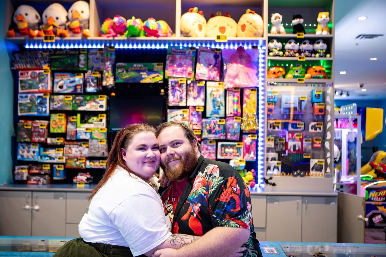 A couple embraces in front of colorful toy store shelves filled with plush animals and games.