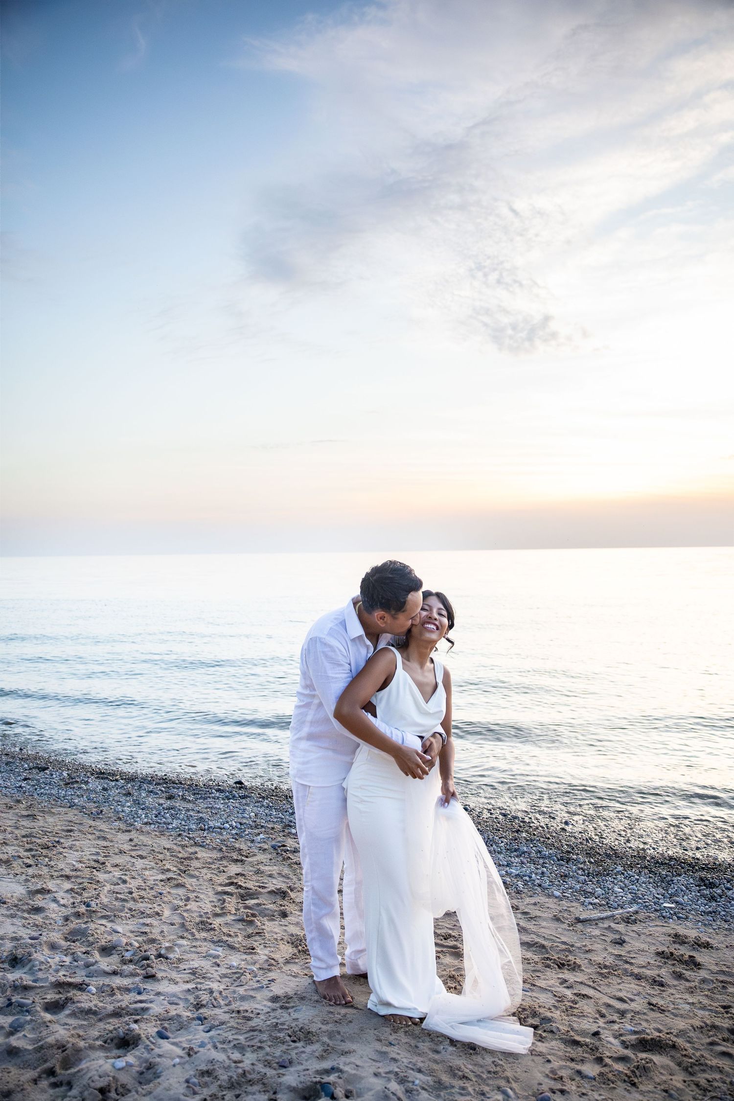 Couple in white attire sharing a romantic kiss on a sandy beach at sunset with calm ocean waves behind them.
