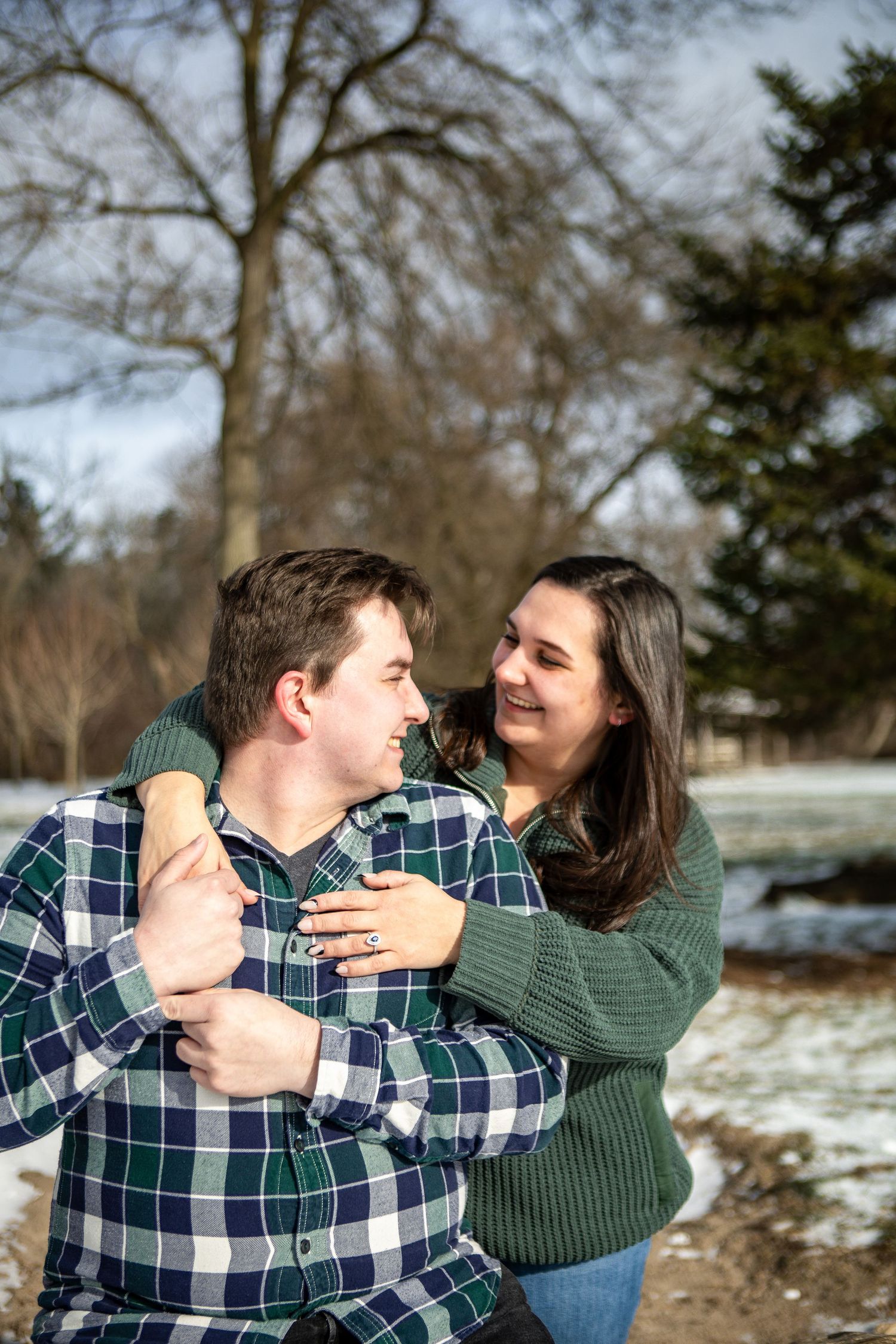 Couple embracing and smiling at each other outdoors in winter, wearing green and plaid shirts with snow in background.