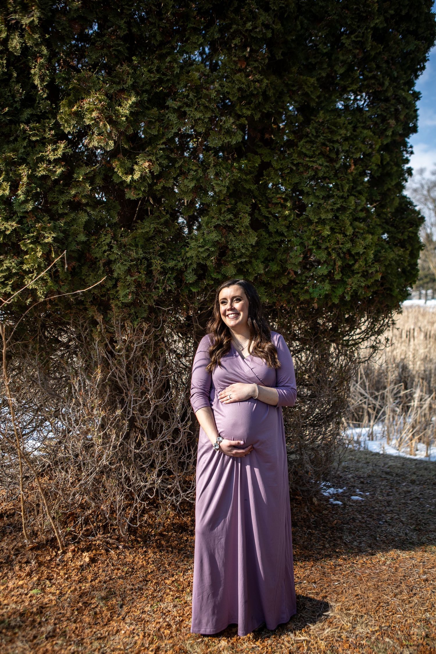 Smiling pregnant woman in purple maxi dress posing outdoors near evergreen tree in winter setting.