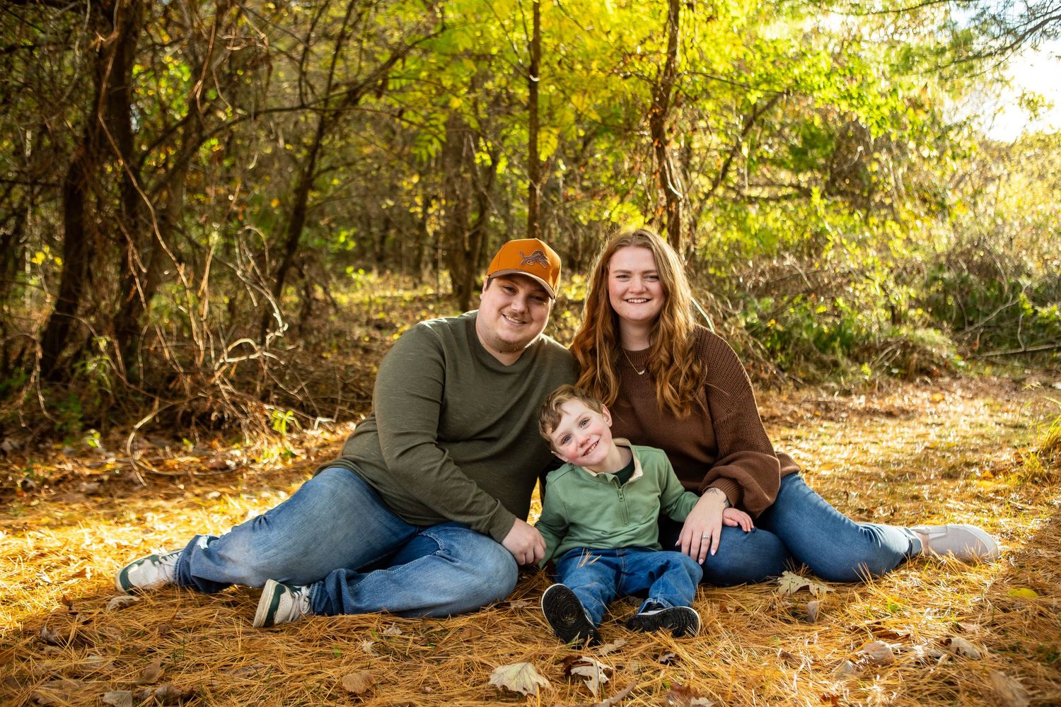 A family poses together on fallen autumn leaves in a sunlit forest setting during an outdoor photo session.