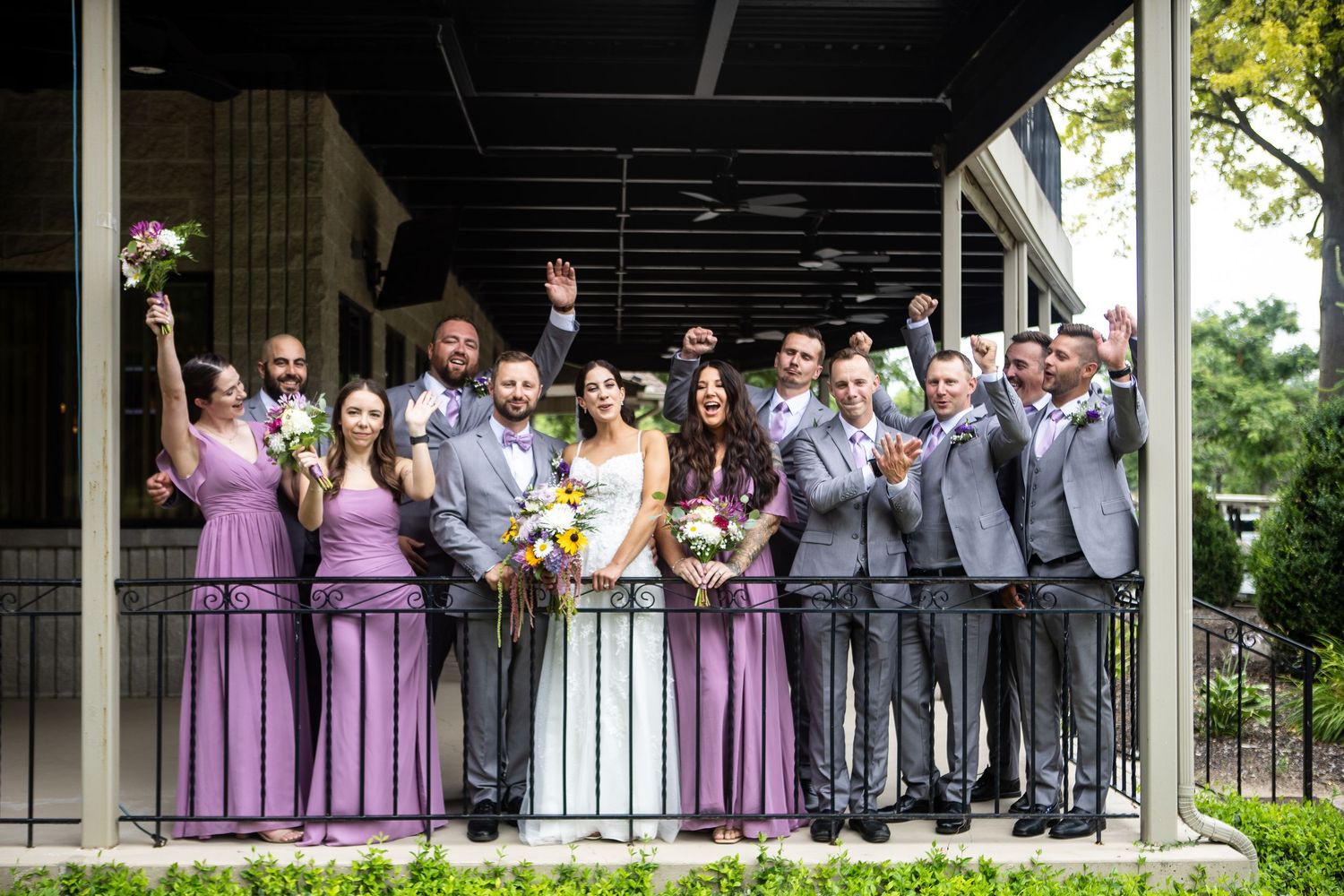 Wedding party celebrates on balcony, bridesmaids in lavender gowns and groomsmen in gray suits cheering with bride and groom.