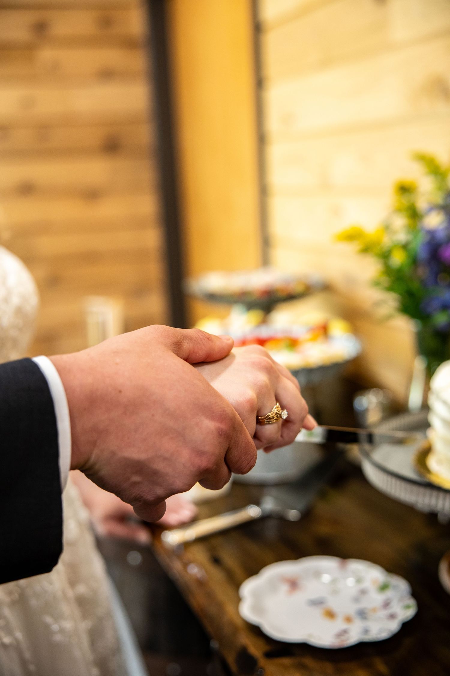 Bride and groom's hands joined on a knife while cutting their wedding cake at a rustic reception with wood walls and fresh flowers.