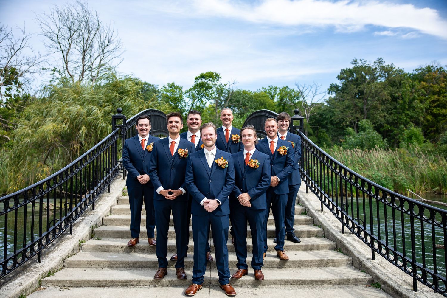 Groomsmen in navy suits with orange boutonnieres pose on stone steps of an arched bridge surrounded by lush green trees at a wedding.