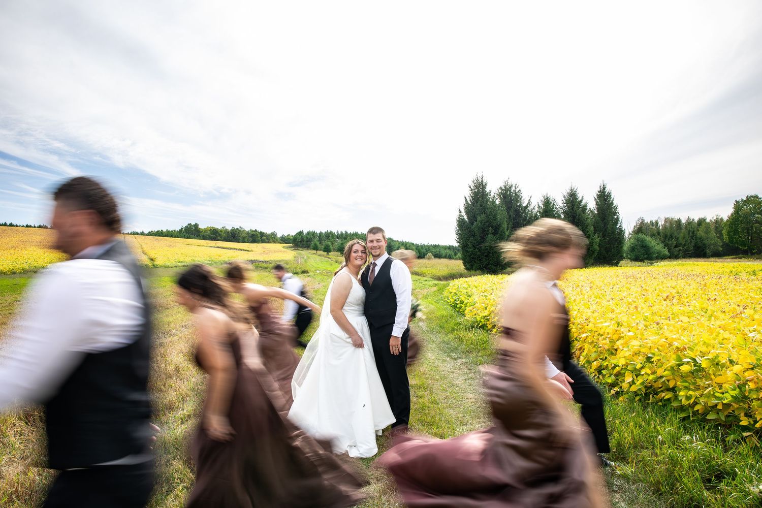 Bride in white gown and groom in vest and tie pose in a field of yellow flowers as blurred wedding guests rush past in motion.