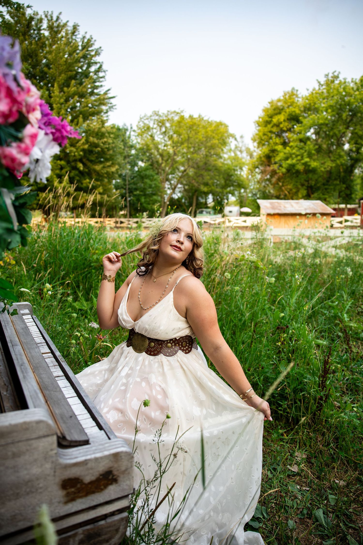 Blonde woman in white boho dress with brown belt poses by vintage piano in lush green field with flowers.