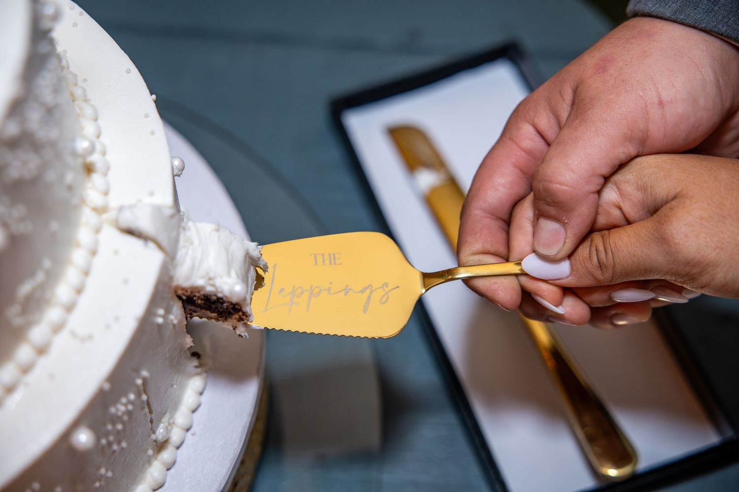 Hands holding a gold engraved cake server cutting a slice from a white pearl-decorated wedding cake, with a cake knife in a gift box nearby.