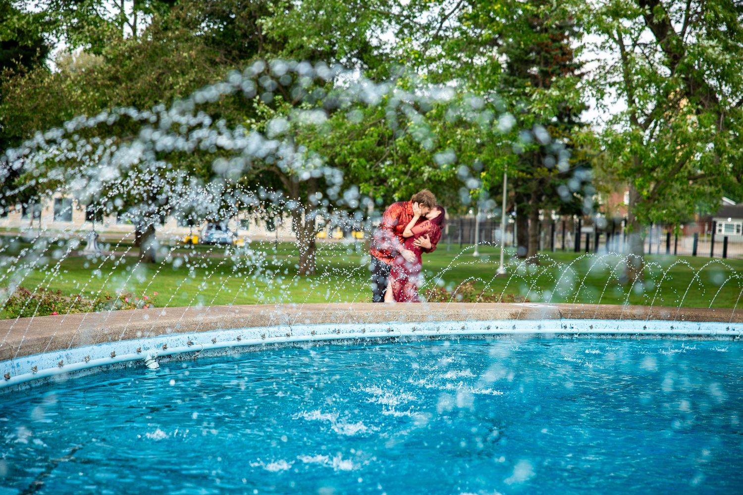 Child playing near a splashing outdoor pool surrounded by green trees on a sunny day.