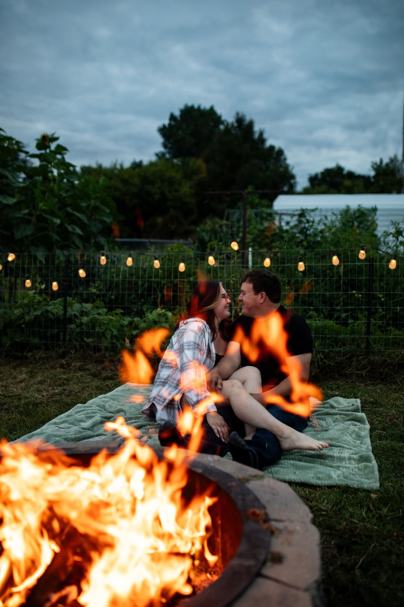 Couple lounging on a blanket by a campfire with string lights creating a romantic outdoor atmosphere.