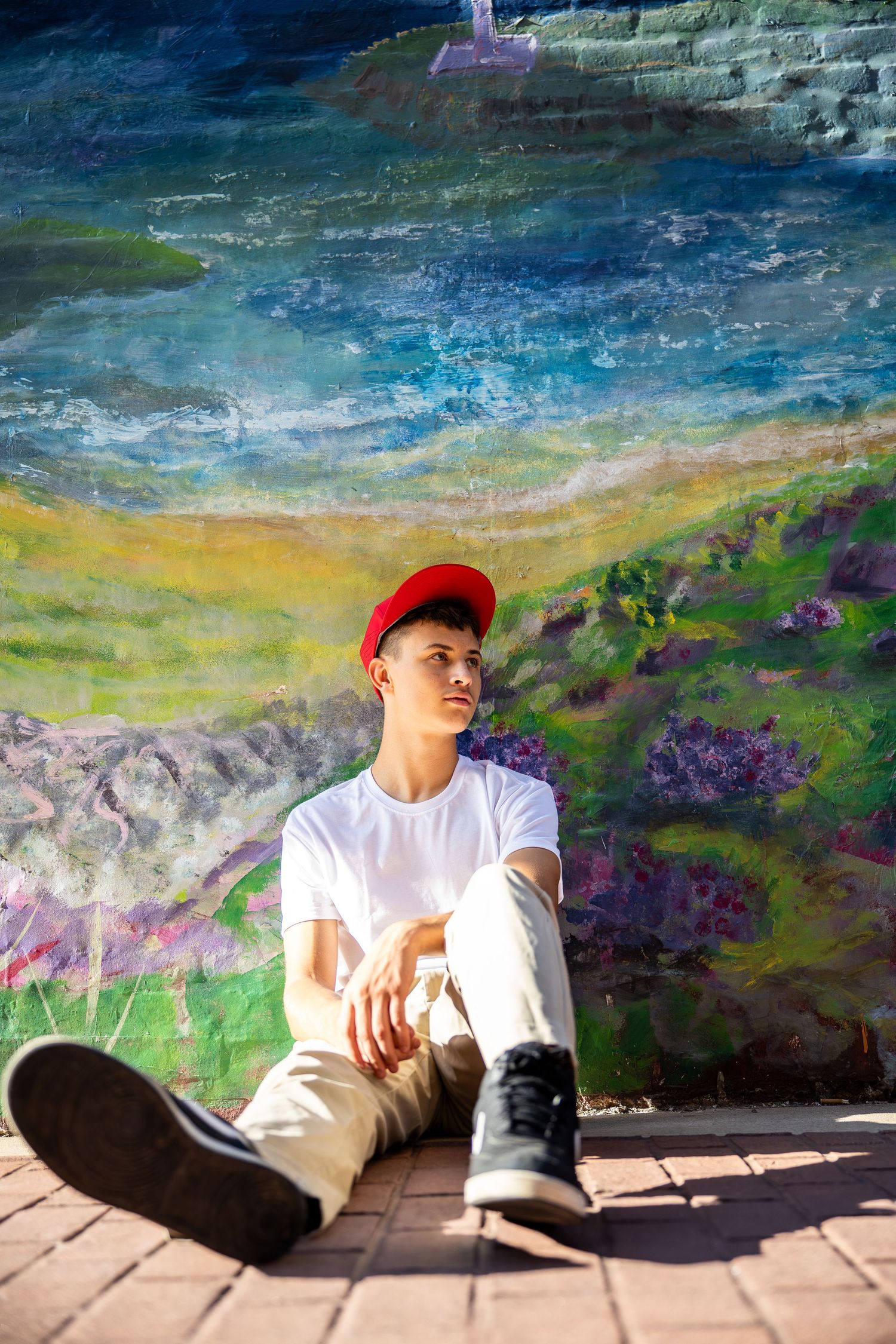 Young man in red cap and white tee sits against colorful mural wall on brick ground.