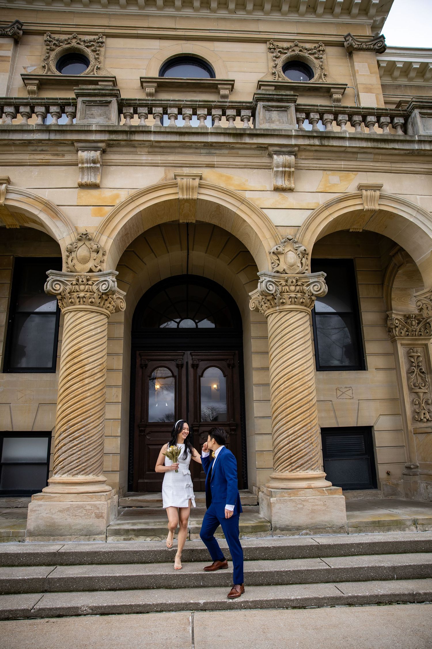 Couple in wedding attire standing on steps of ornate stone building with arched doorway and spiral columns.