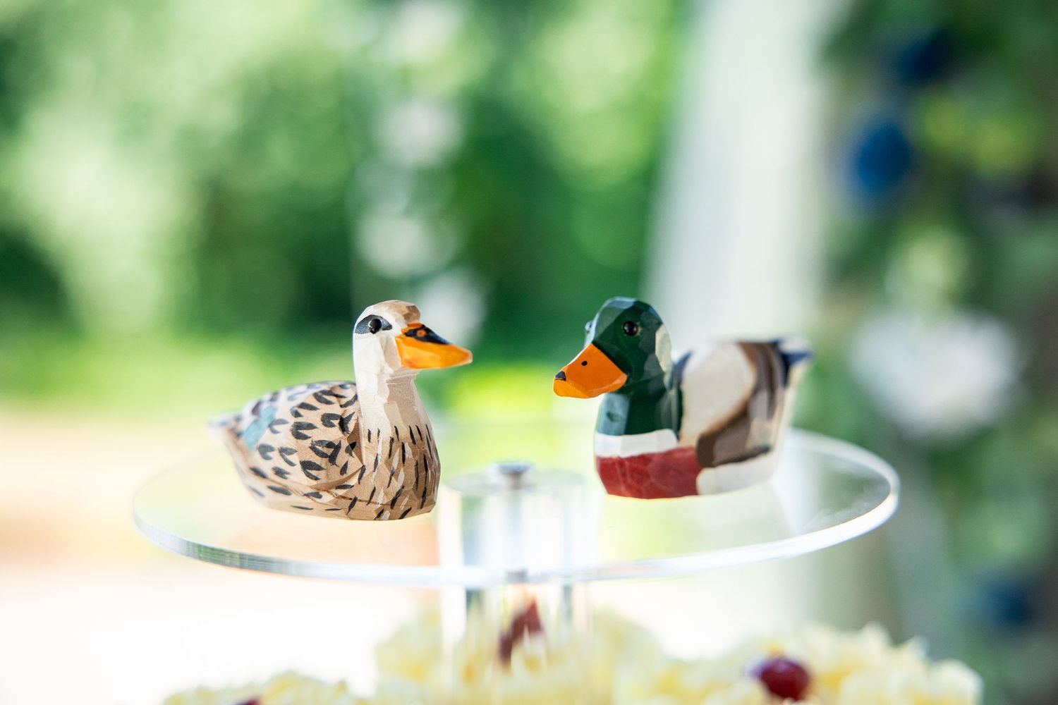 Two decorative mallard duck figurines displayed on a glass stand with a blurred green garden background.