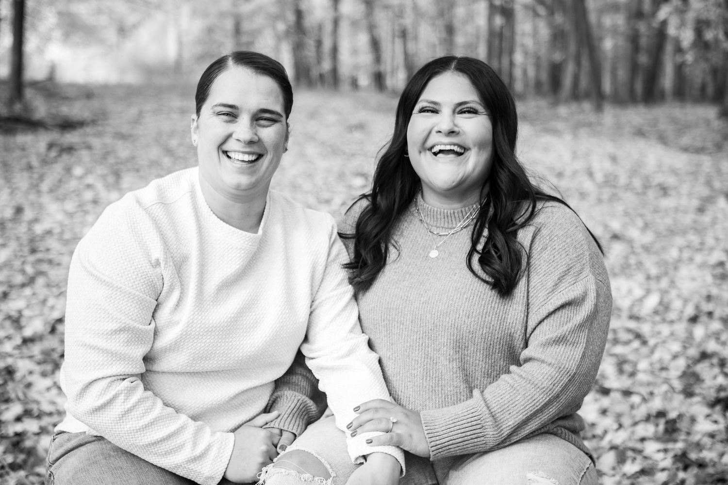 Black and white photo of two smiling people sitting outdoors among fallen leaves, holding hands in a warm autumn setting.