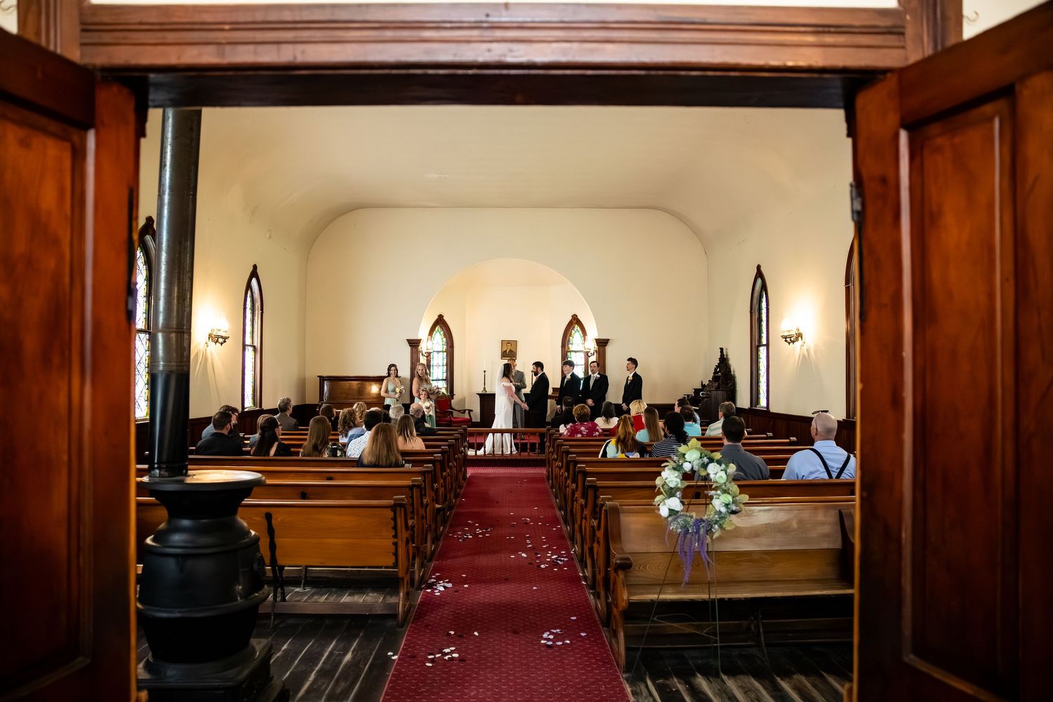 A wedding ceremony inside a charming historic church with red carpet aisle, wooden pews, and arched windows.