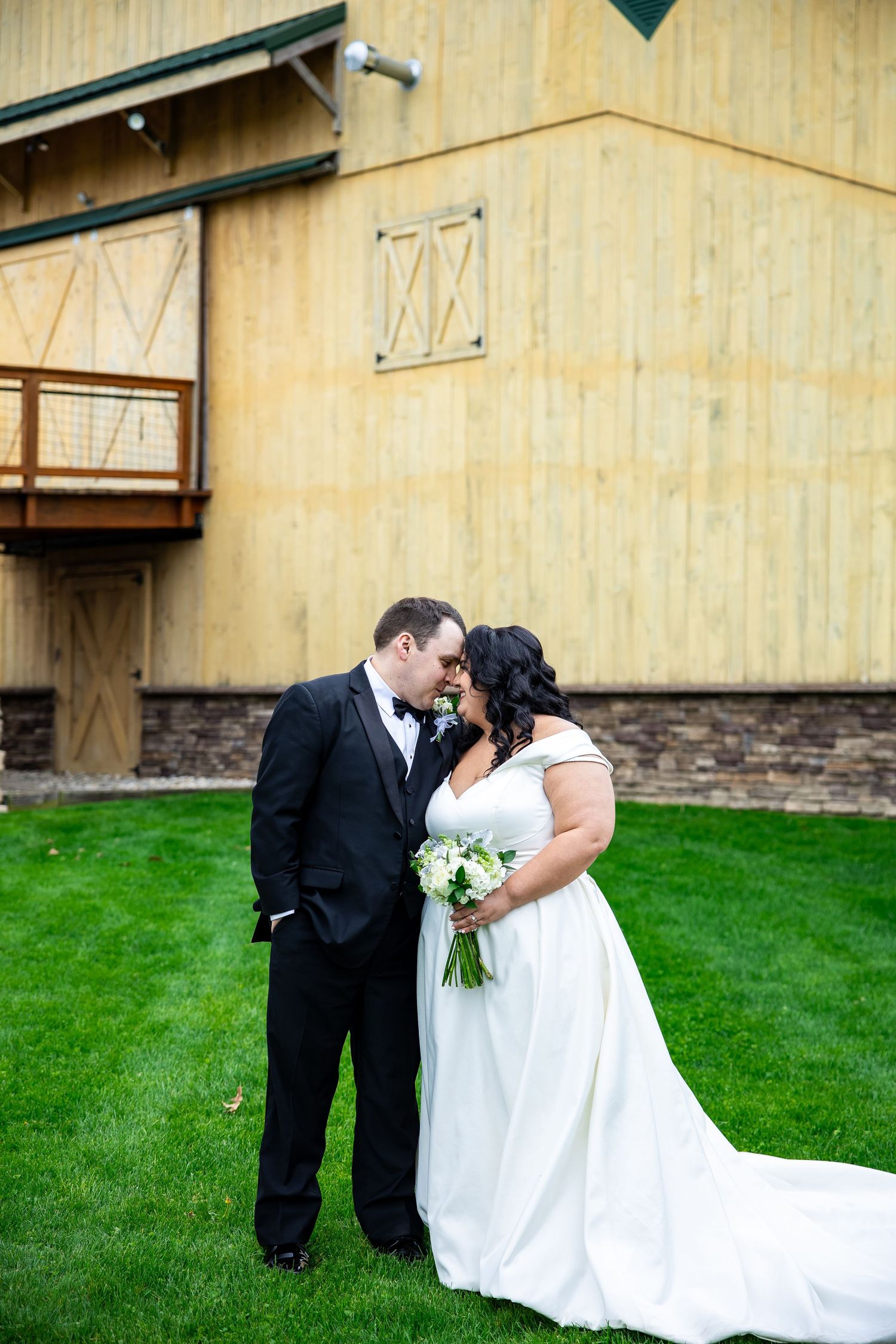 Bride and groom share a romantic moment kissing in front of a rustic barn venue on lush green grass.