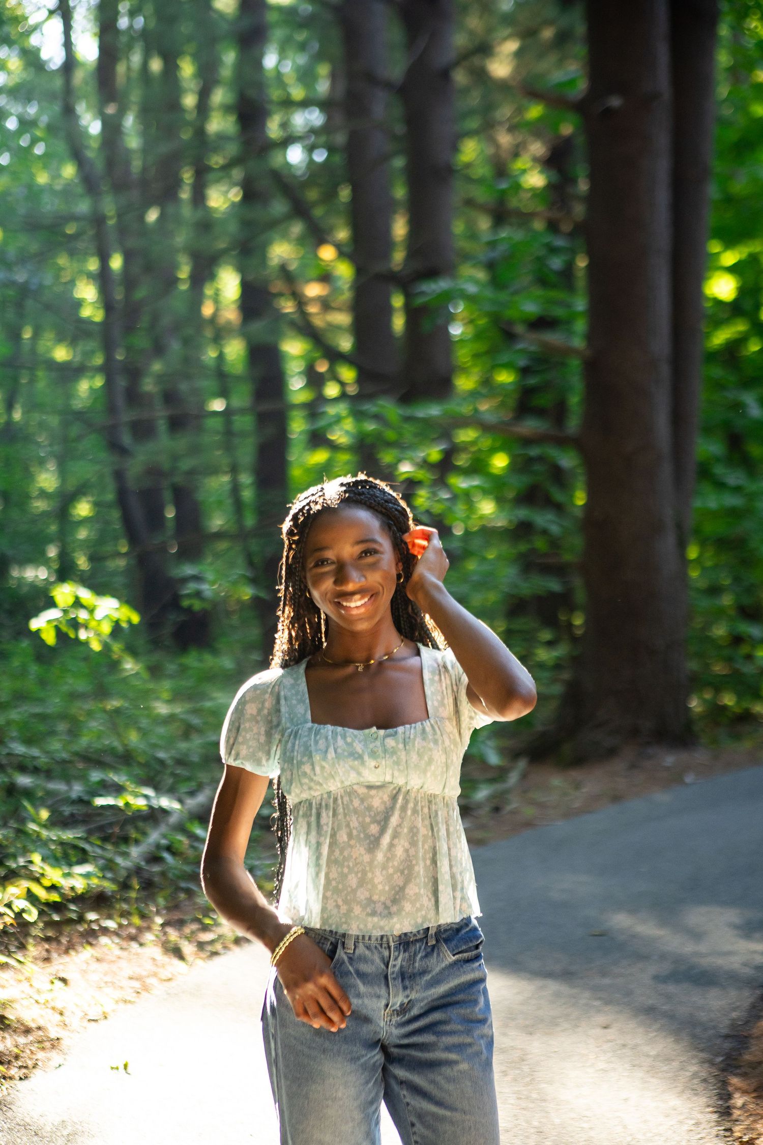 A person wearing a white lace top and jeans poses playfully in a sunlit forest path.